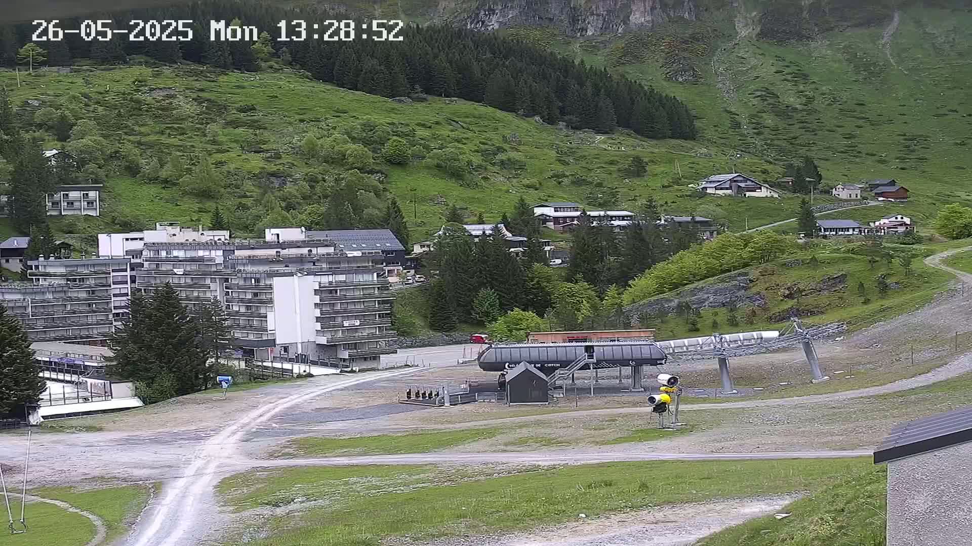 A ski lift base station sits in a mountain valley, with a large apartment building and several smaller buildings visible, under an overcast sky.