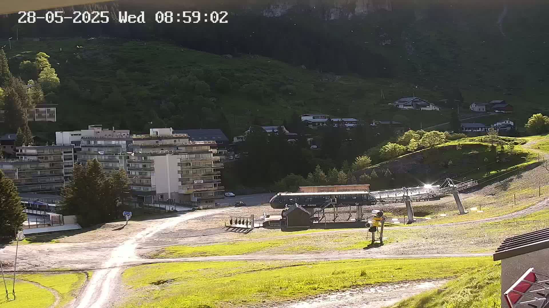 A sunny mountainside scene shows a chairlift, a multi-story building, and several smaller buildings nestled among green hills and trees.