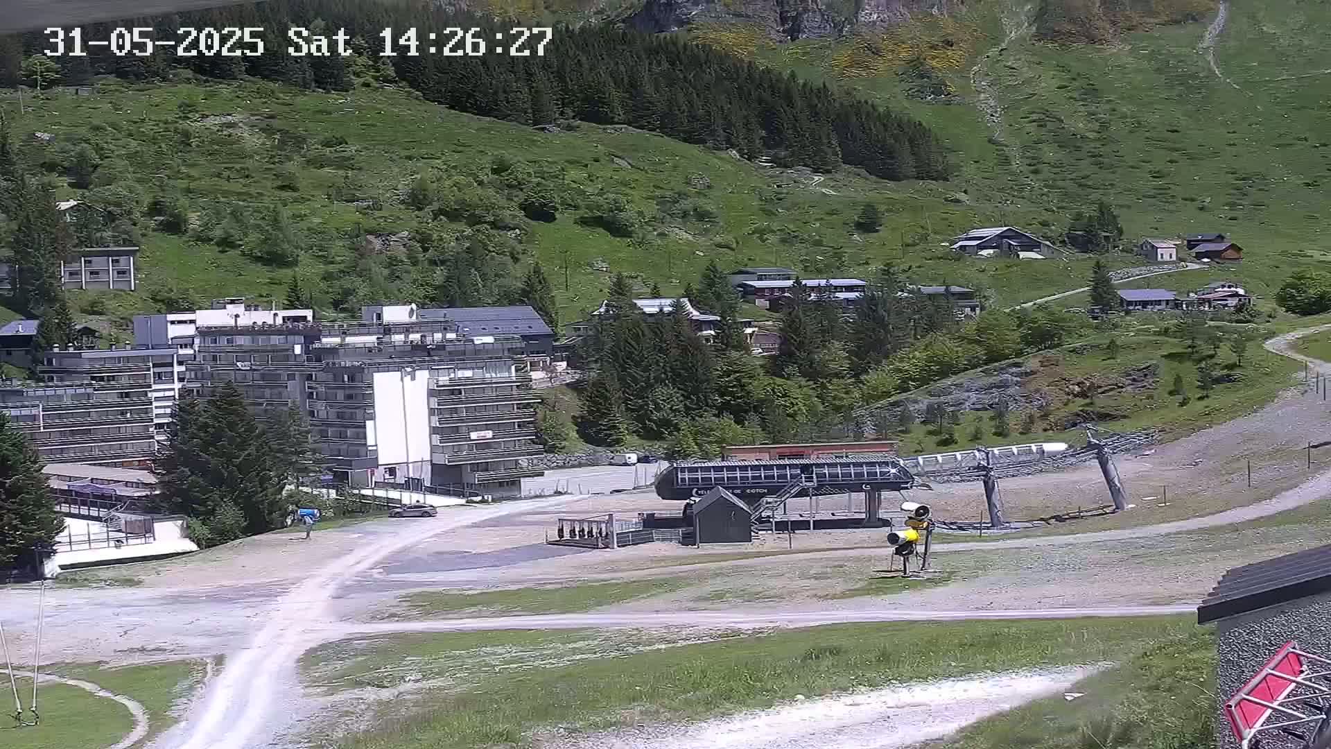 A ski lift base station and several buildings are situated on a mountainside on a sunny day.