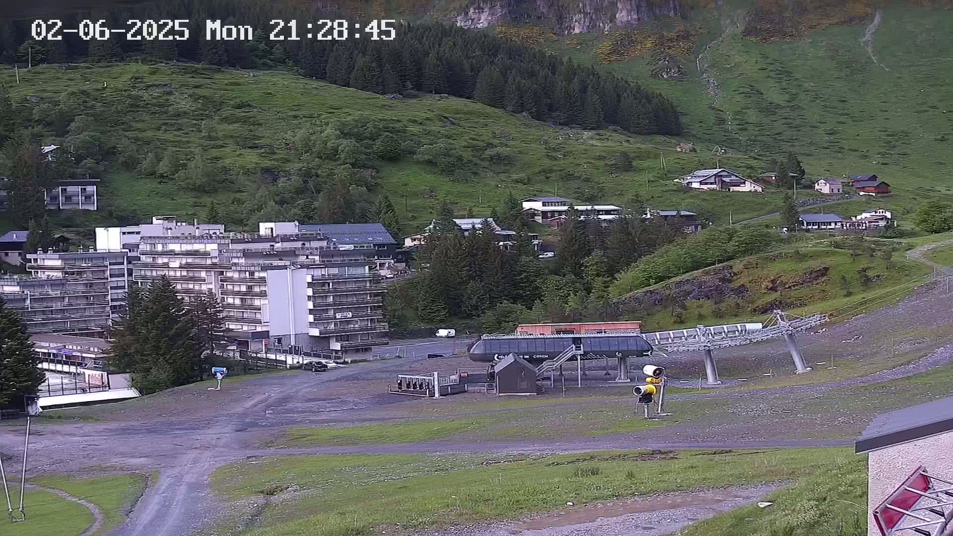 A multi-story building and several smaller buildings are situated on a hillside with a chairlift and a mostly barren area in the foreground, under an overcast sky.
