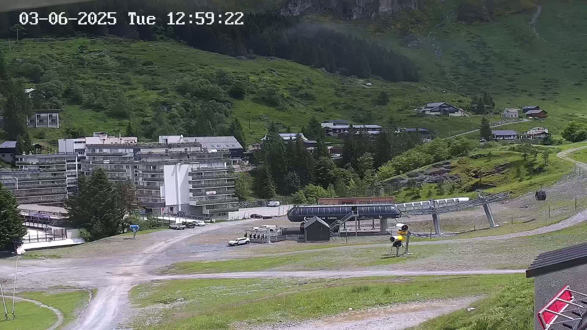 A mountainside village with a large hotel, chairlift, and several smaller buildings is visible on a partly cloudy day.