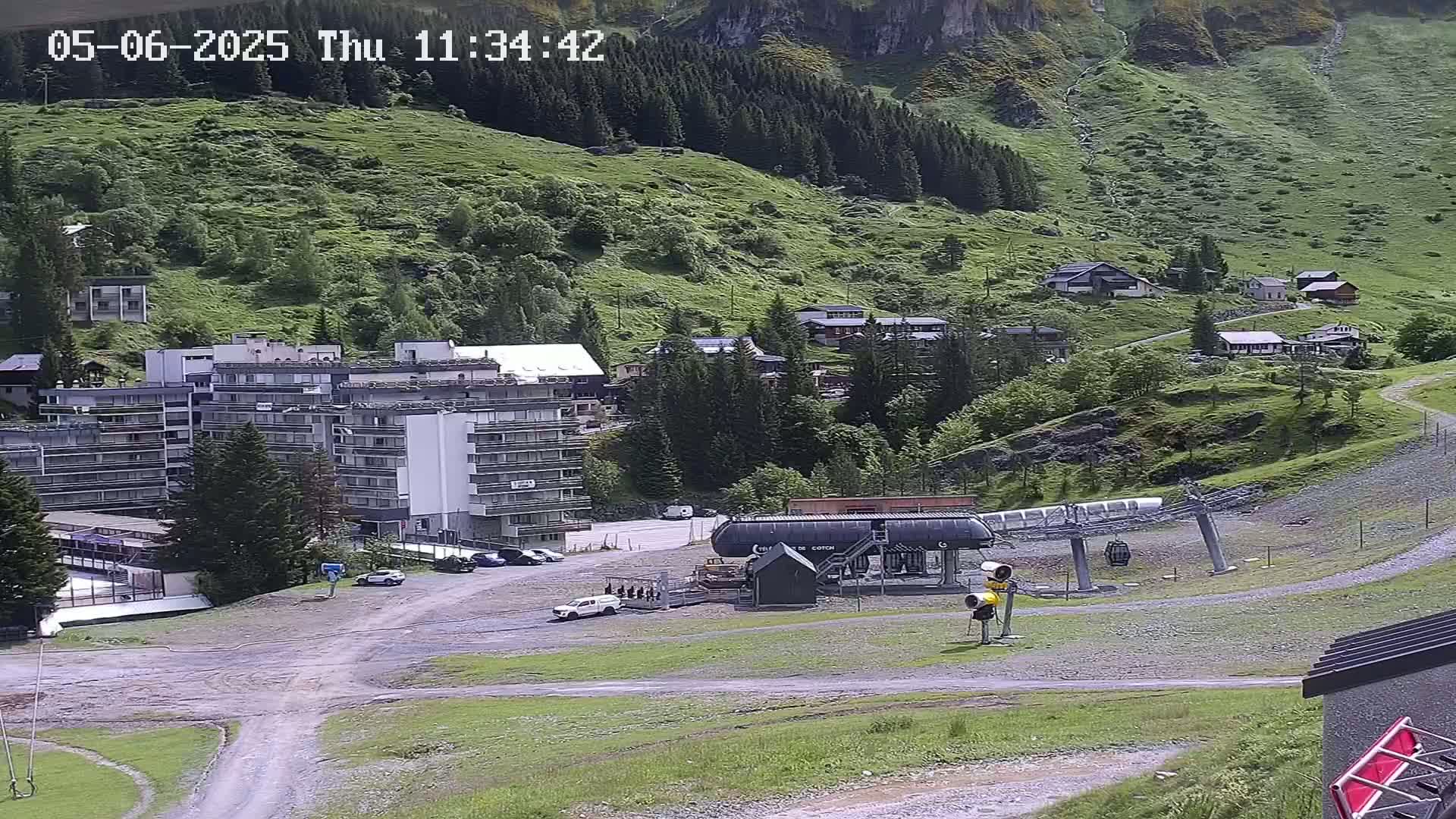 A ski lift base station and several buildings sit on a grassy hillside, with a mountain in the background under a mostly clear sky.