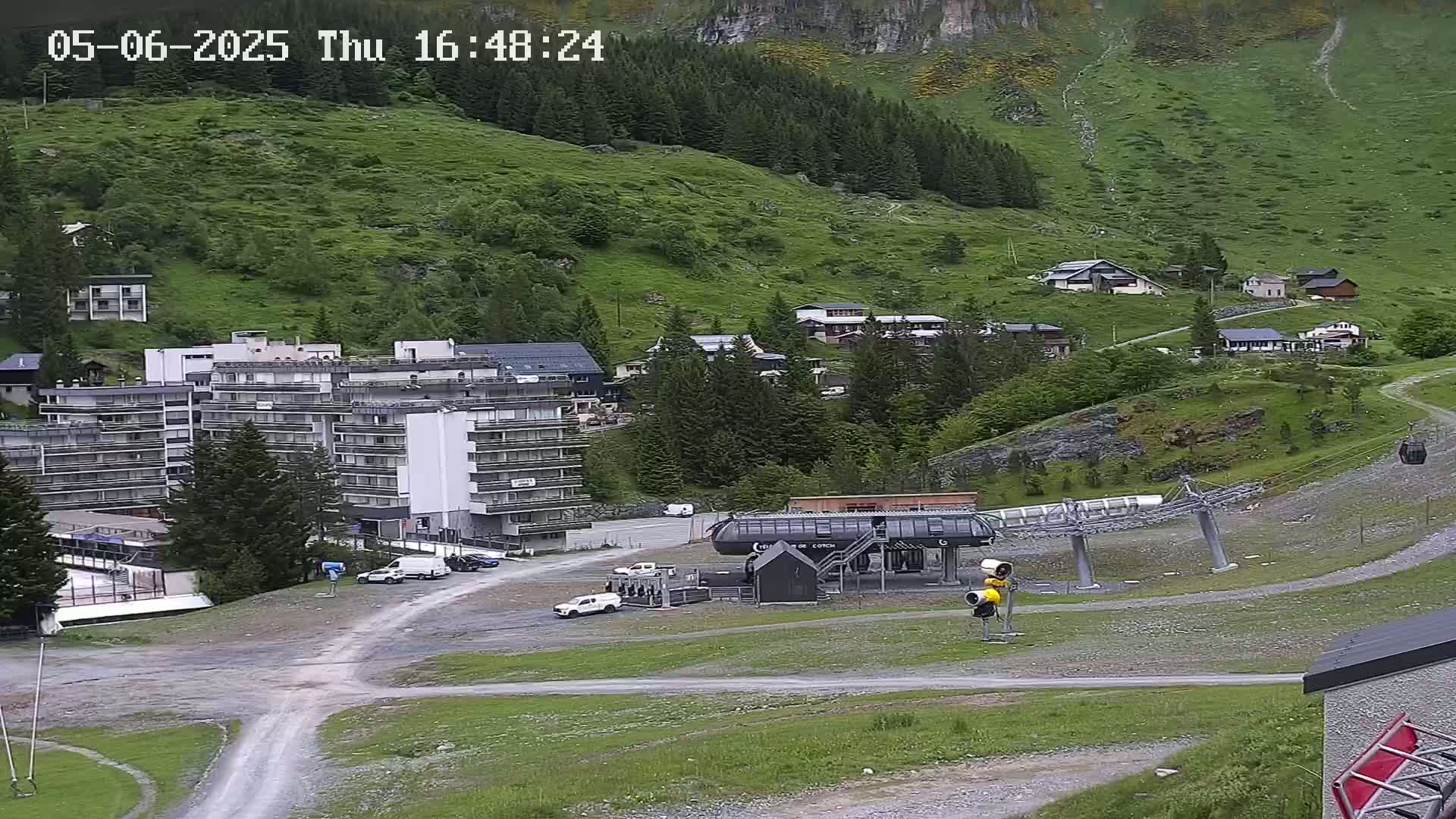 A mountainside village with several buildings, a chairlift, and cars in a parking lot is visible under a mostly cloudy sky.