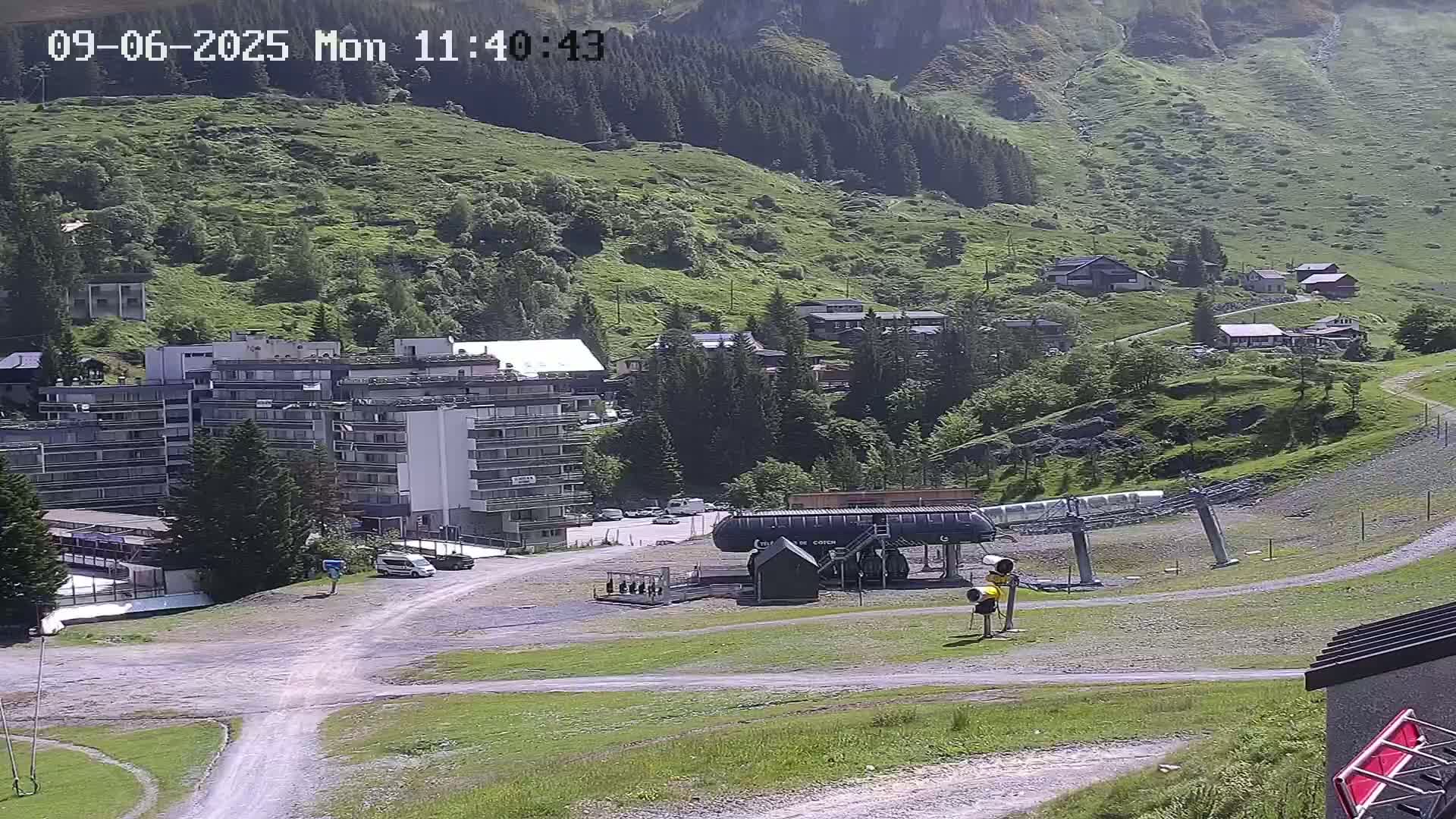A sunny view of a mountainside village with a large apartment building, a chairlift, and several smaller buildings nestled amongst green hills and trees.
