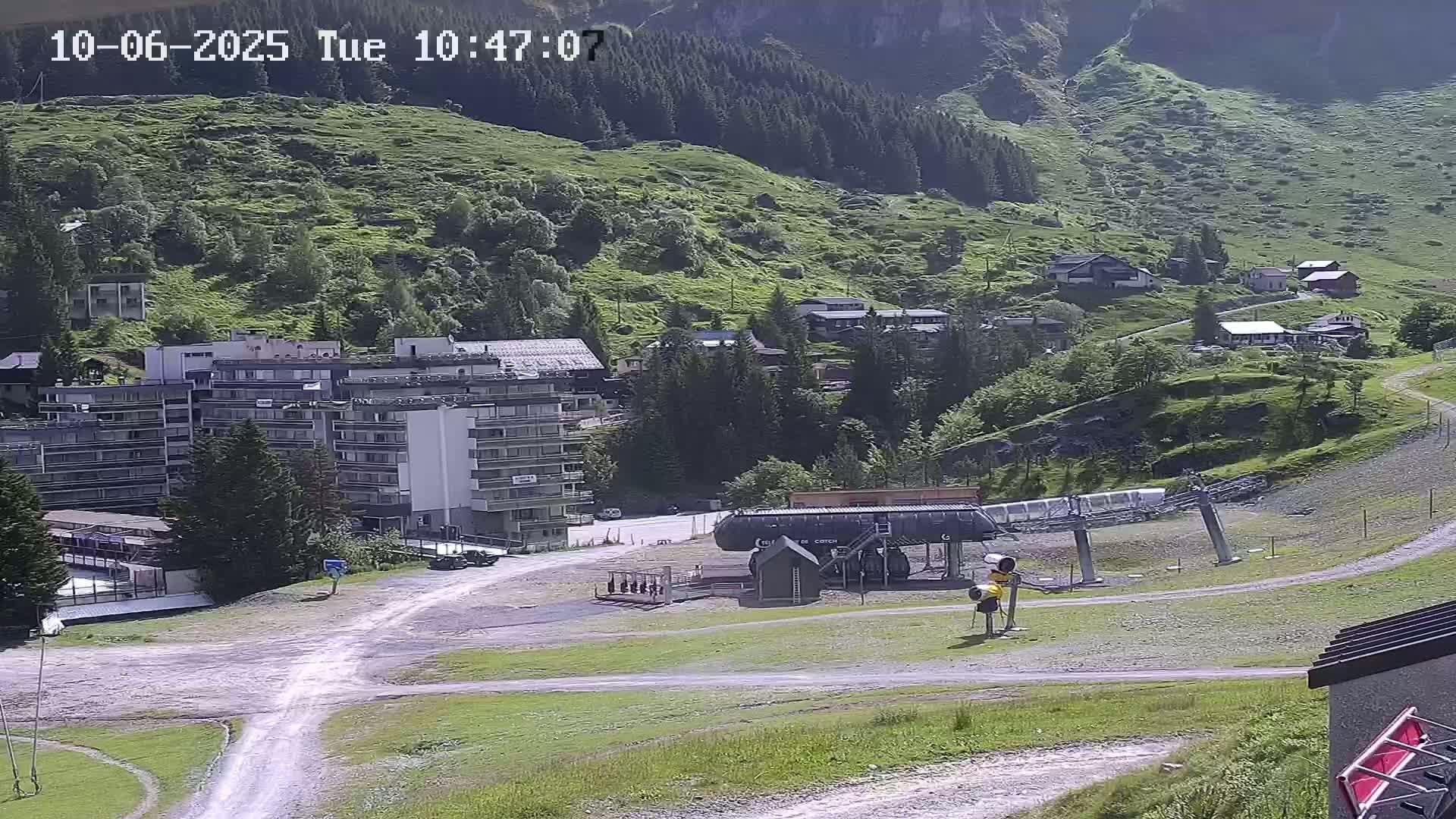 A mountainside village with a large apartment building, a chairlift, and several smaller buildings is visible on a sunny day.