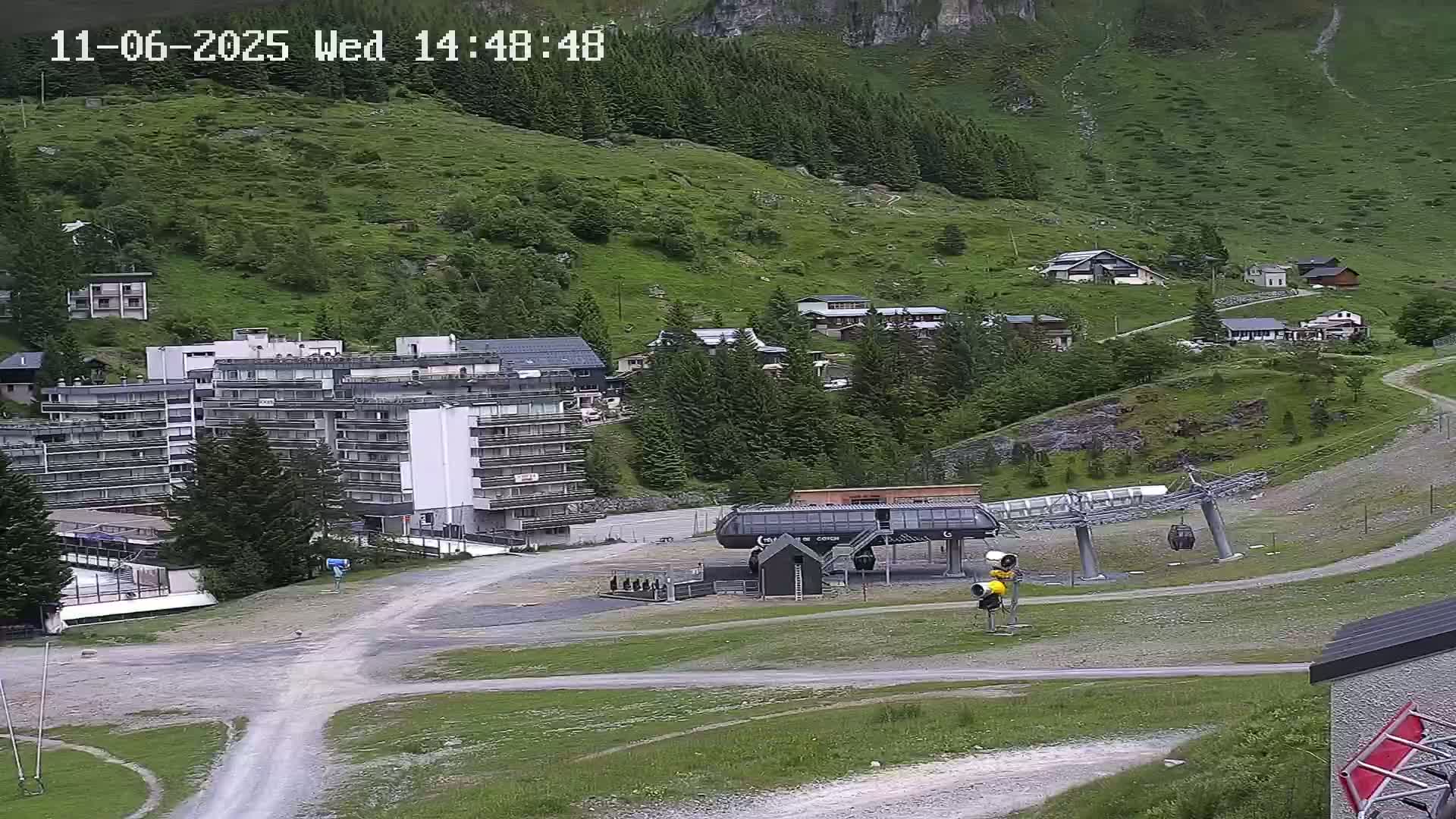 A ski lift base station sits in a mountain valley town on an overcast day, with several buildings and green hills visible.