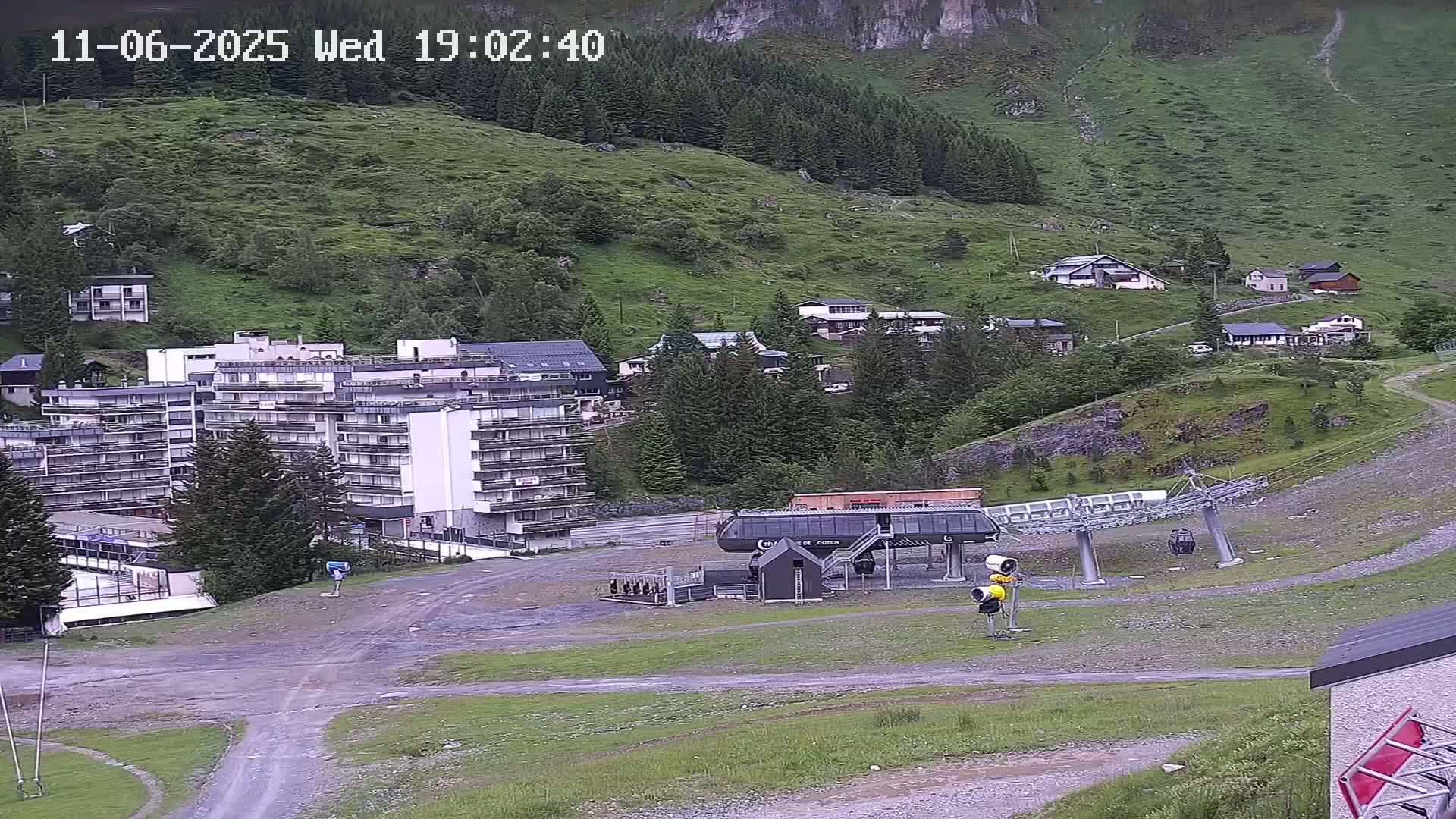 A chairlift base station sits in a gravel area next to a multi-story building, with a hillside of green vegetation and small buildings in the background under overcast skies.