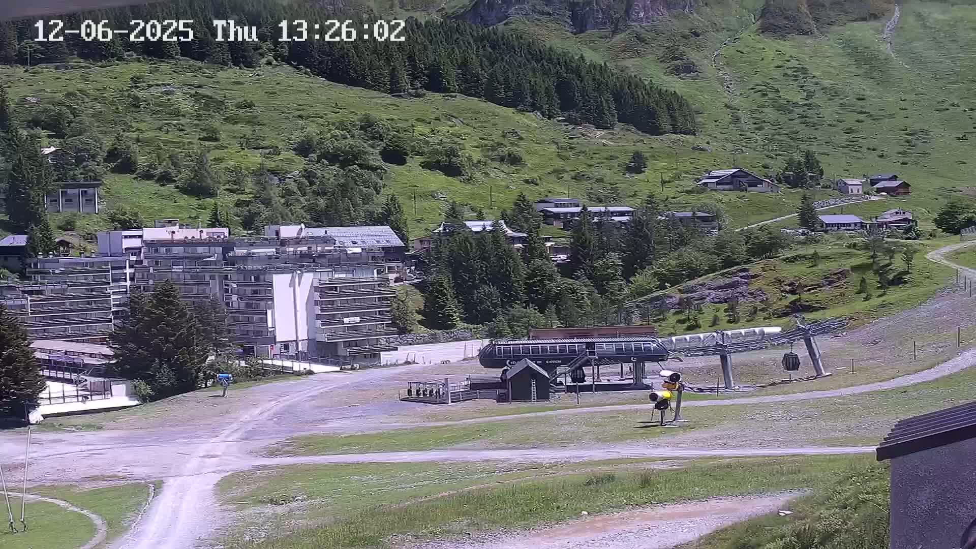 A ski lift base station sits in a mountain valley on a sunny day, near several buildings and green hillsides.