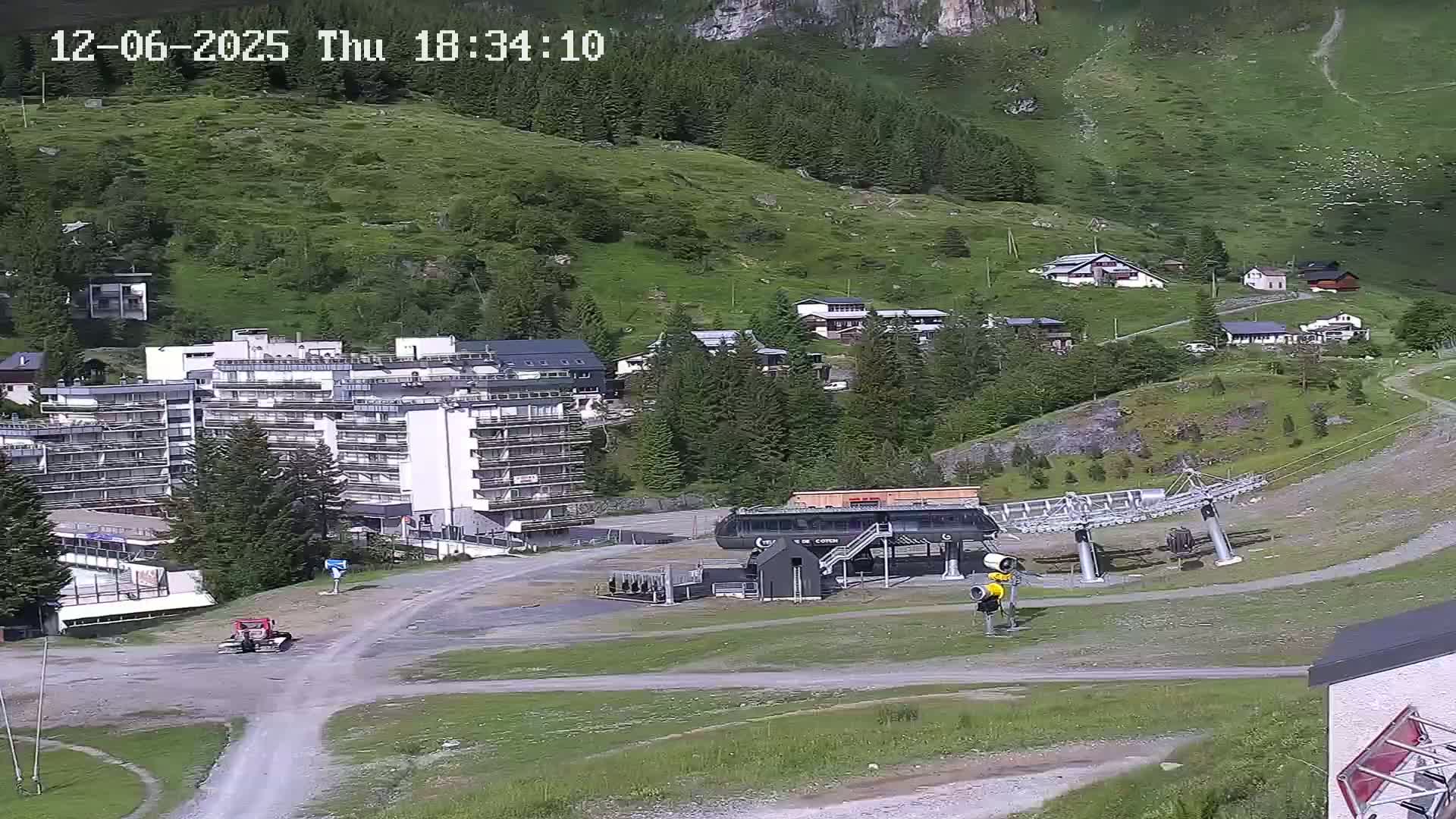 A sunny mountainside village with a large apartment building, a chairlift, and several smaller buildings is visible from a high vantage point.