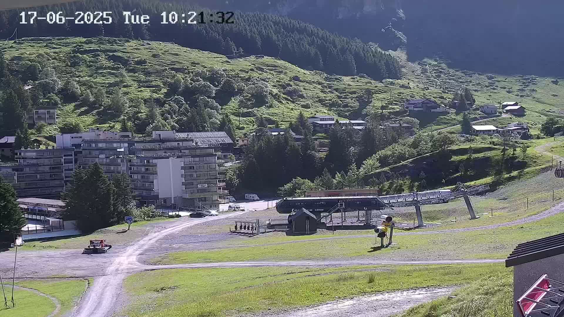 A sunny mountainside view shows a ski lift base station, several buildings, and a small vehicle near a dirt road.