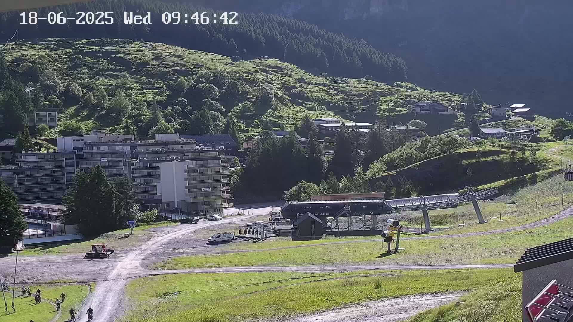 A sunny mountainside village scene shows a chairlift, several buildings, including a large apartment building, a few vehicles, and several people biking on a path.
