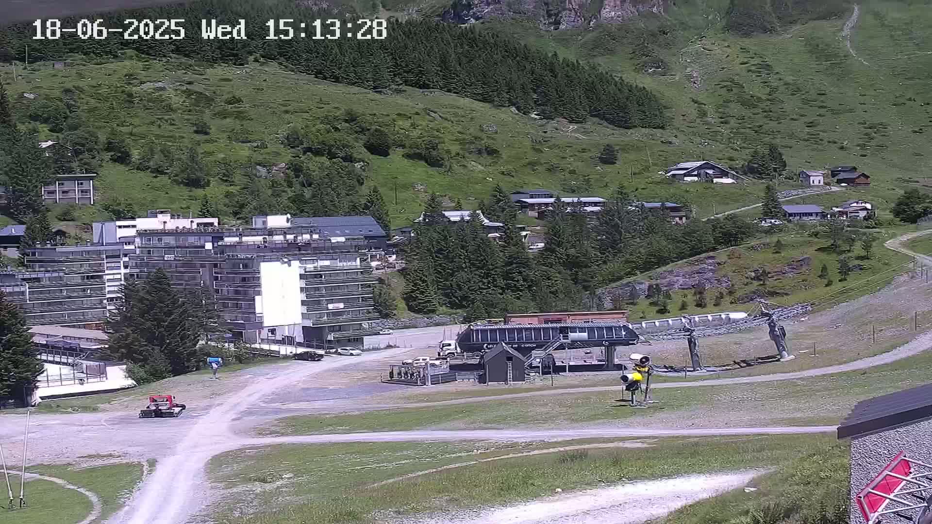 A ski lift base station sits in a sunny mountain valley beside several buildings and a grassy hillside with sparse trees.