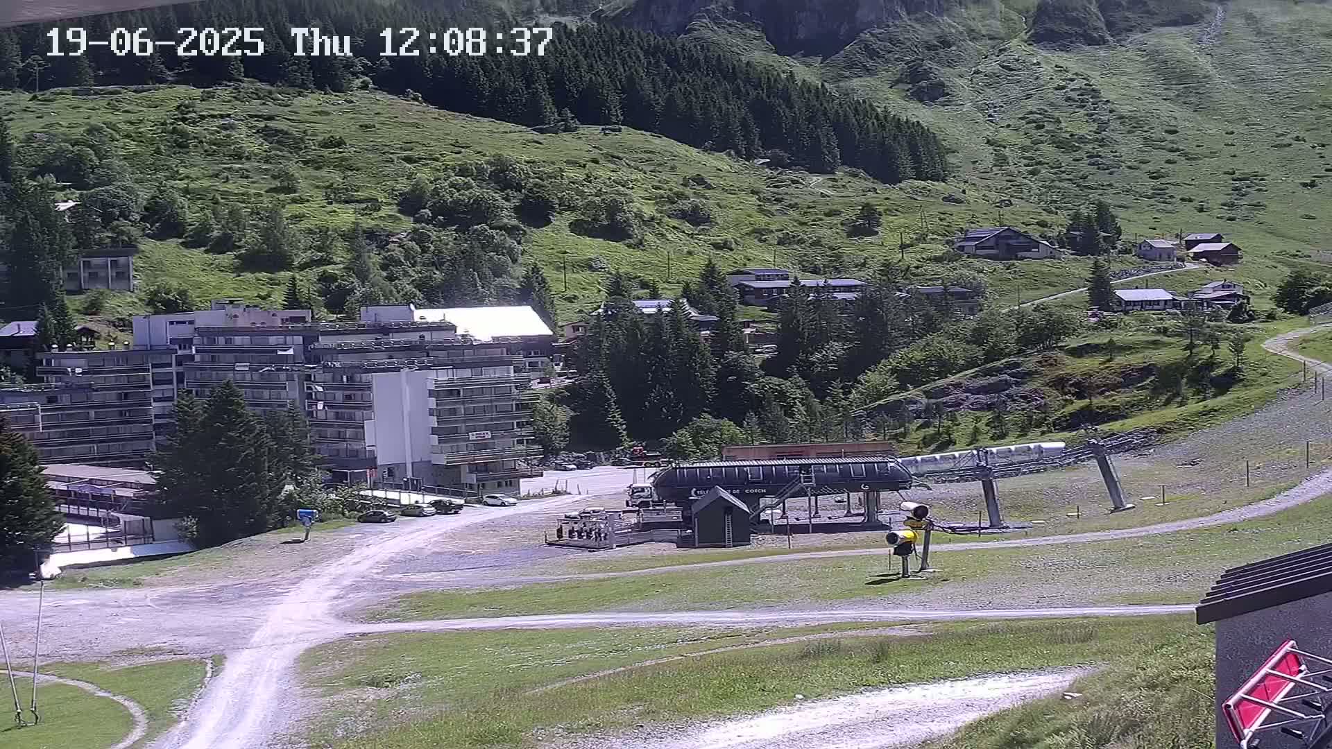 A mountainside village with a multi-story building, a chairlift, and a gravel road is visible on a sunny day.