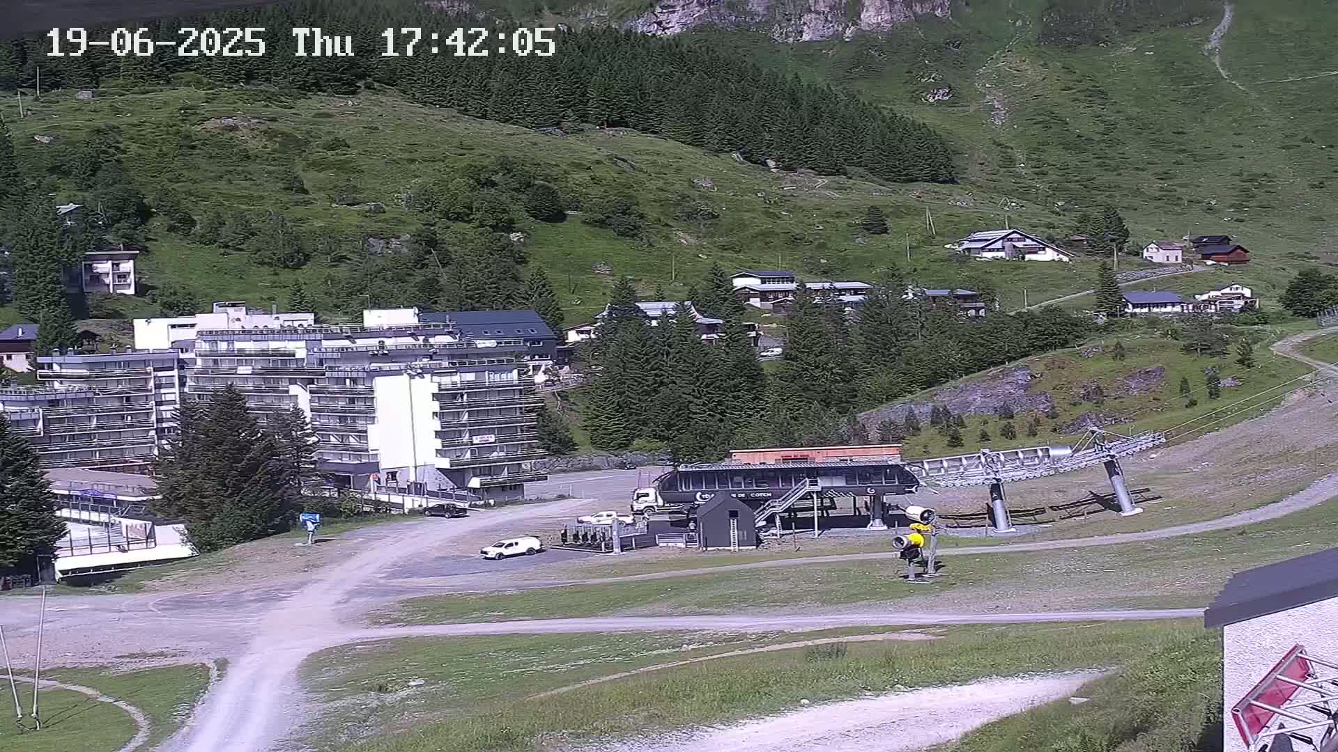 A mountainside village with a large apartment building, a chairlift base station, and several smaller buildings, all under a sunny sky.
