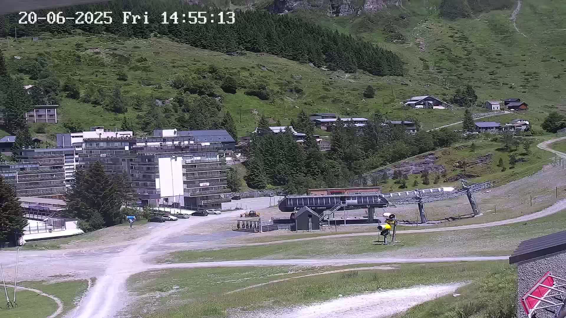 A ski lift base station sits in a sunny mountain valley next to a multi-story building and several smaller buildings, surrounded by green hillsides and coniferous trees.