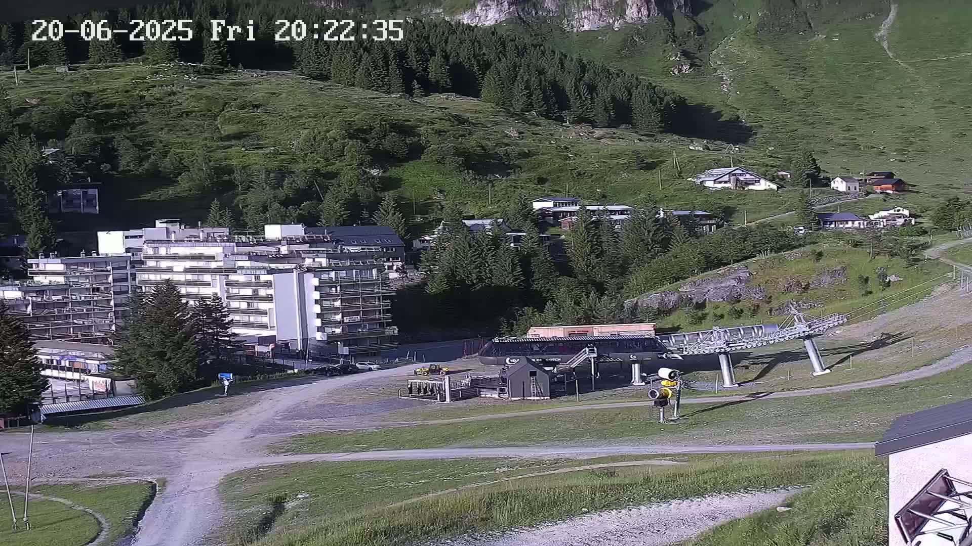A large apartment building and several smaller buildings are situated on a mountainside amongst evergreen trees under sunny conditions, with a chairlift visible in the foreground.