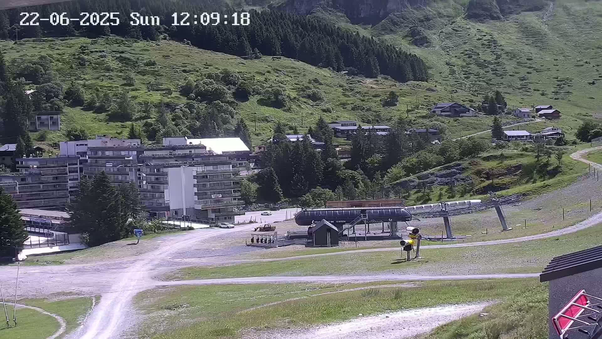 A mountainside village with a large apartment building, a ski lift, and several smaller buildings, under sunny skies.