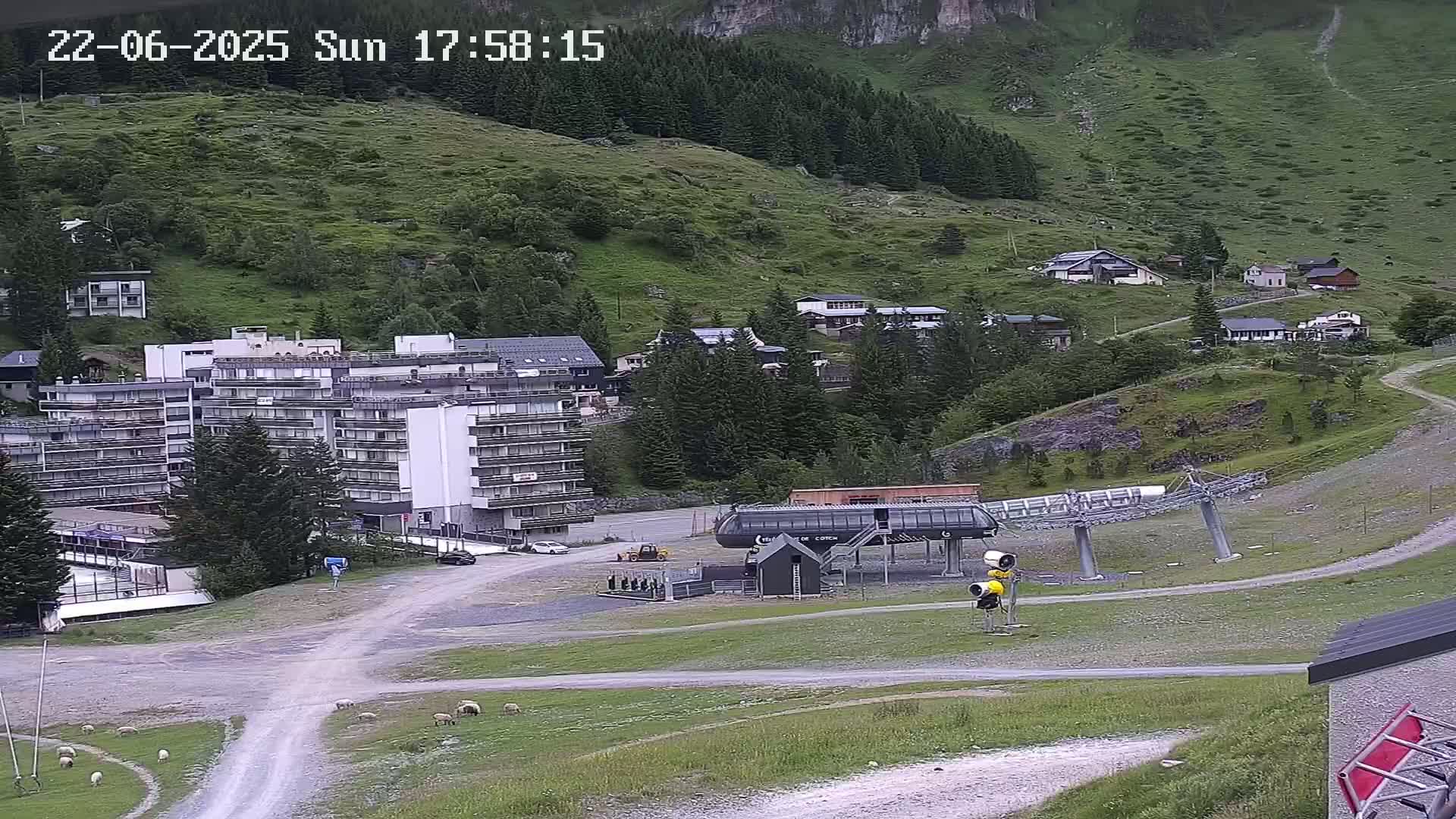 A mountainside village with a large apartment building, a chairlift, and sheep grazing on grassy areas under partly cloudy skies.
