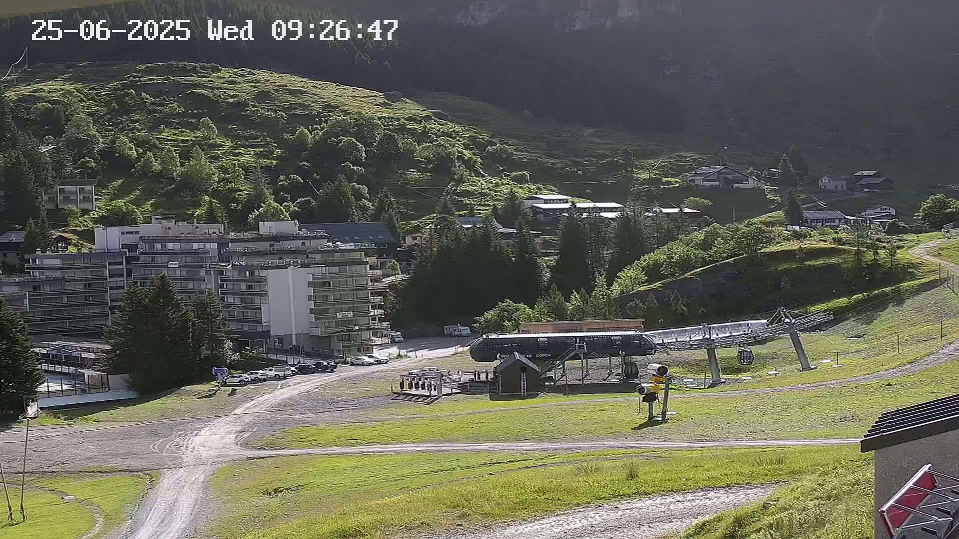 A sunny mountainside view shows a chairlift base station, a multi-story building, several smaller buildings and green hills.