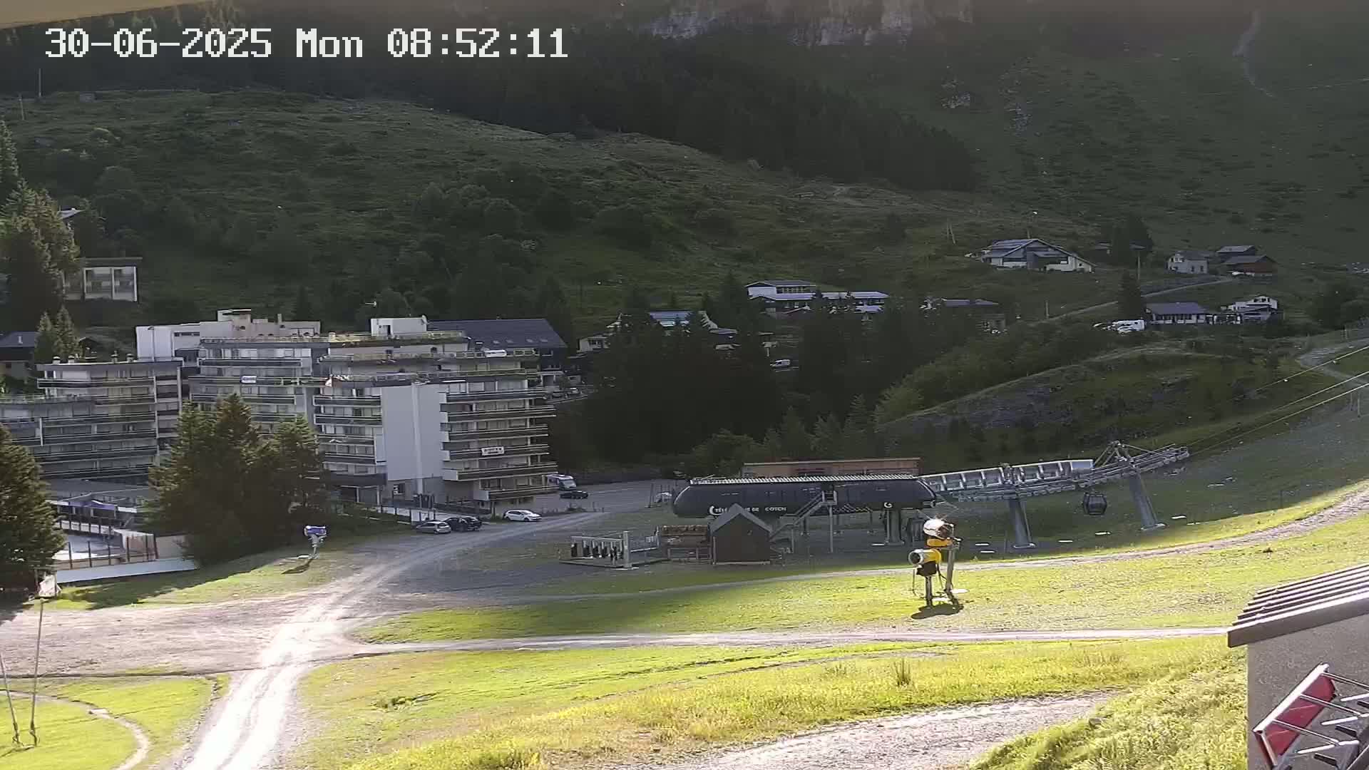 A sunny mountainside view shows a ski lift, several buildings, and a grassy area.