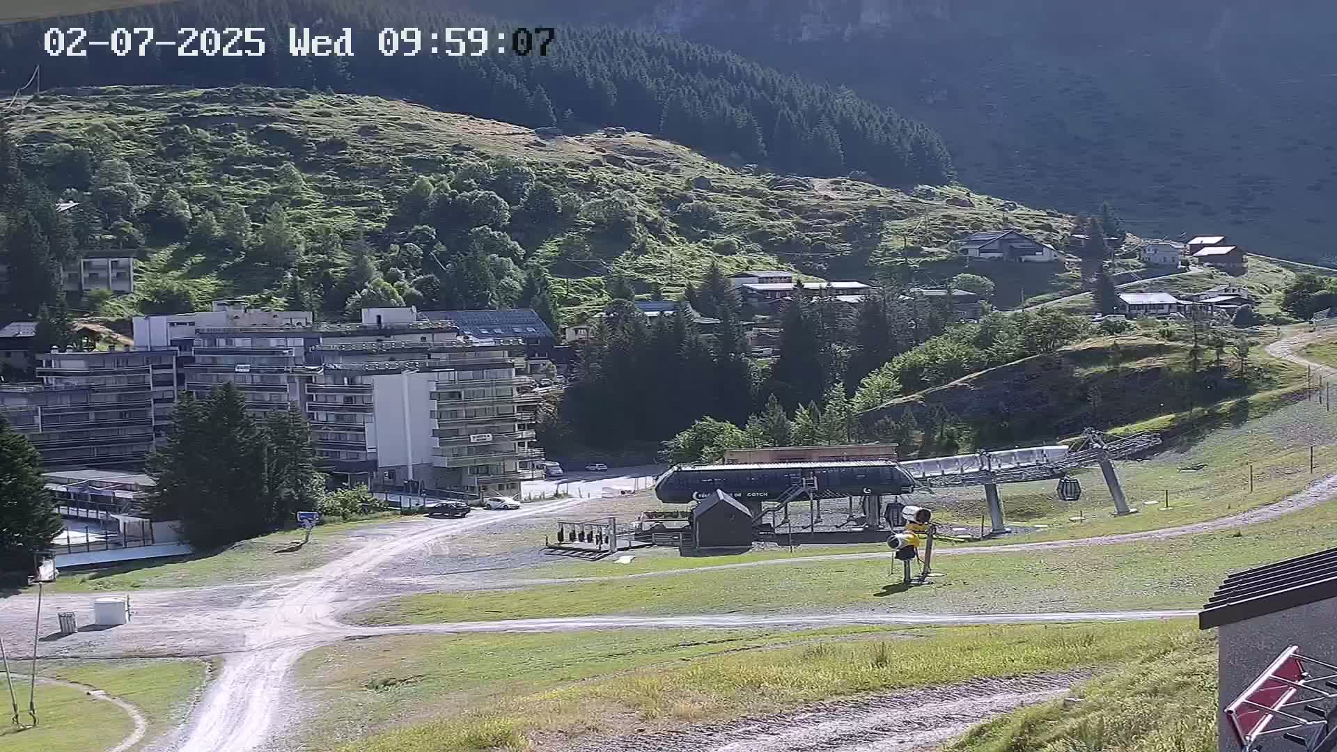 A mountainside village is visible, featuring a multi-story building, a chairlift, and a dirt road under sunny conditions.