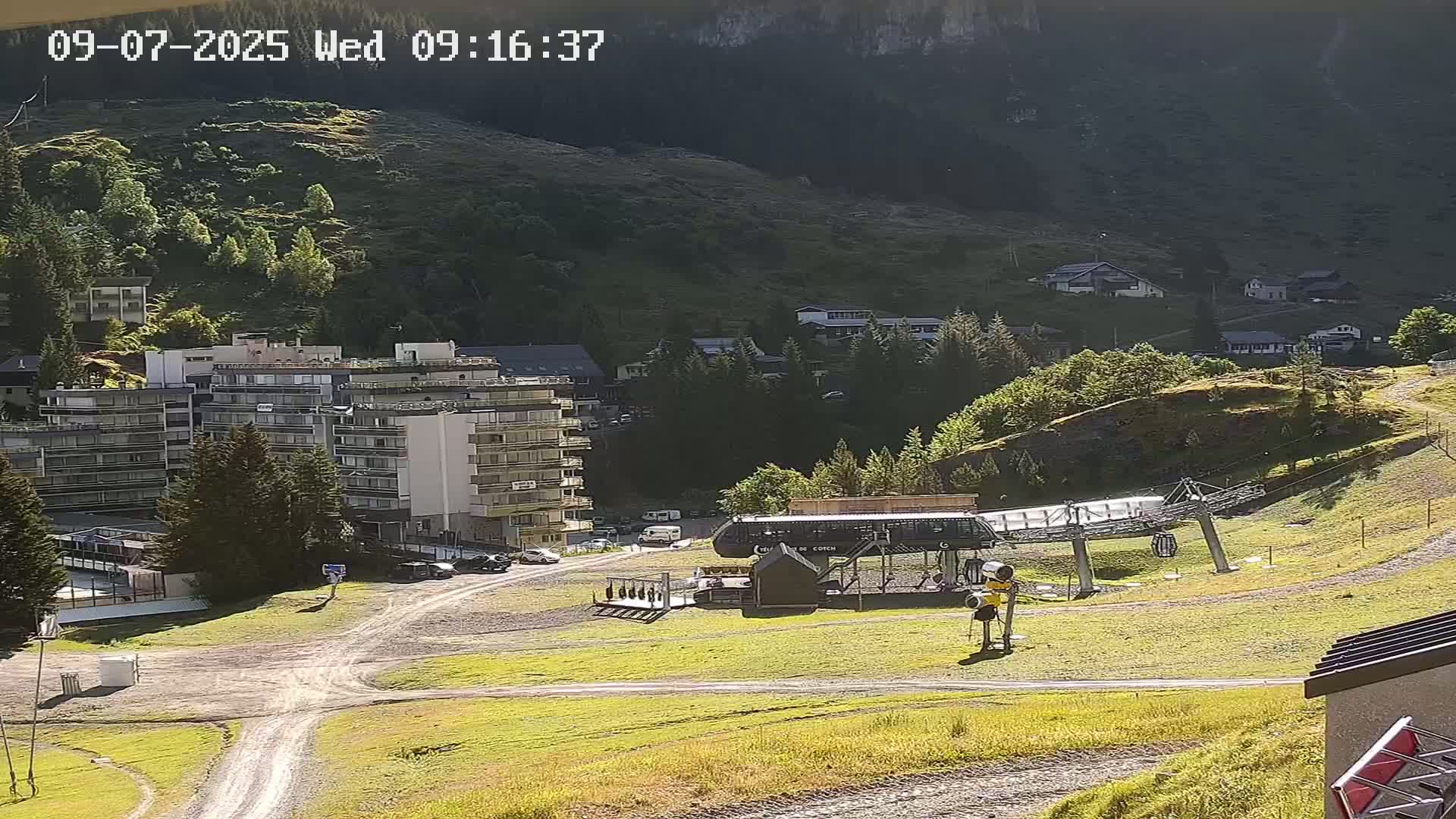 A sunny mountainside view shows a multi-story building, a chairlift, and several smaller buildings nestled among green hills and trees.