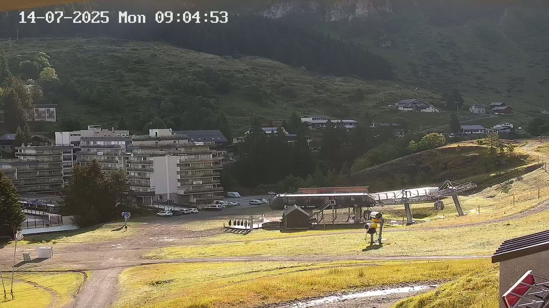 A sunny mountainside view shows a multi-story building, several smaller houses, a chairlift, and grassy areas.