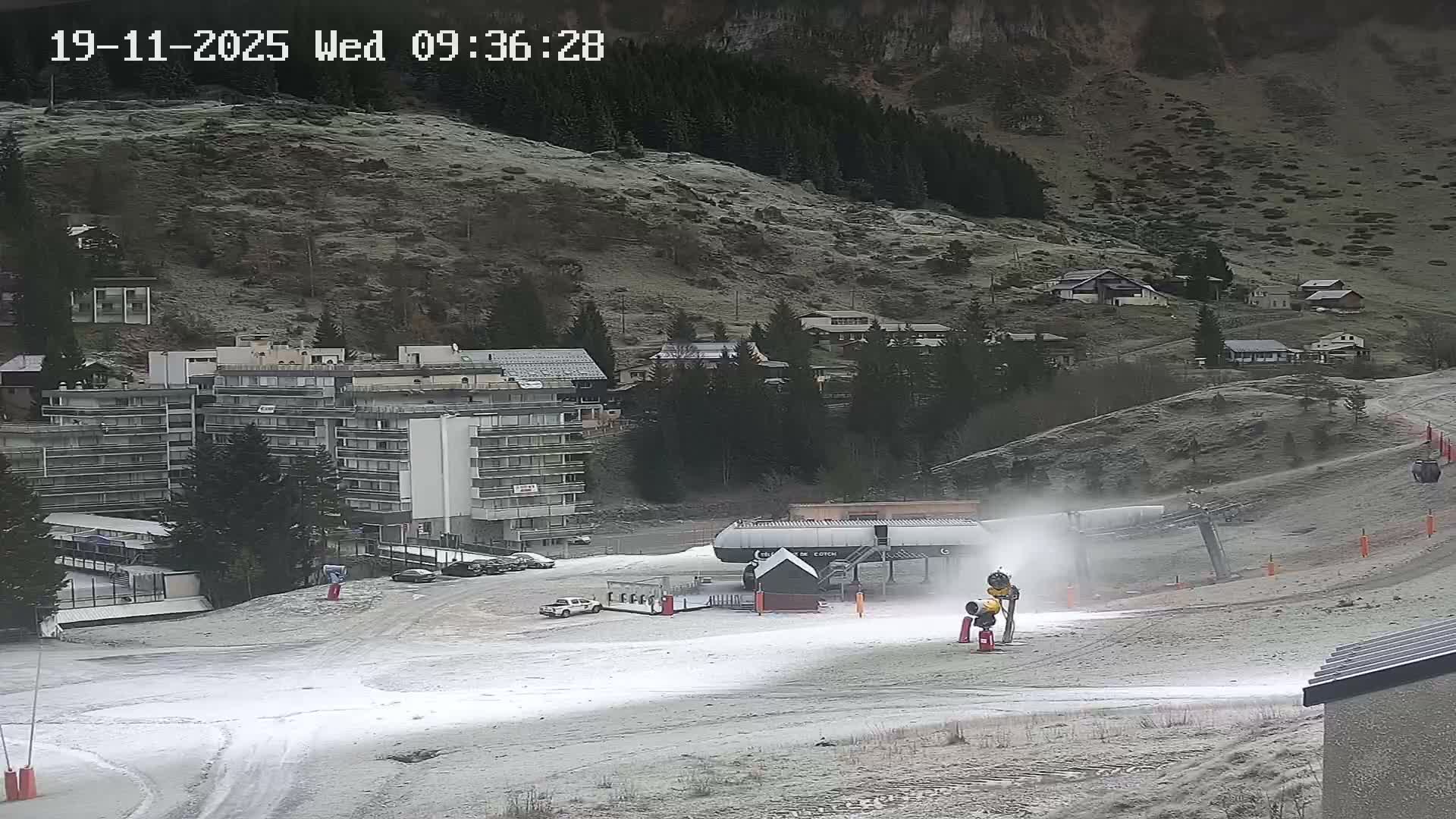 A wintry mountain landscape features a snow cannon actively making snow next to a ski lift station on a partially snow-covered slope, with multiple buildings and frosty, sparsely vegetated hillsides in the background under cold, overcast conditions.