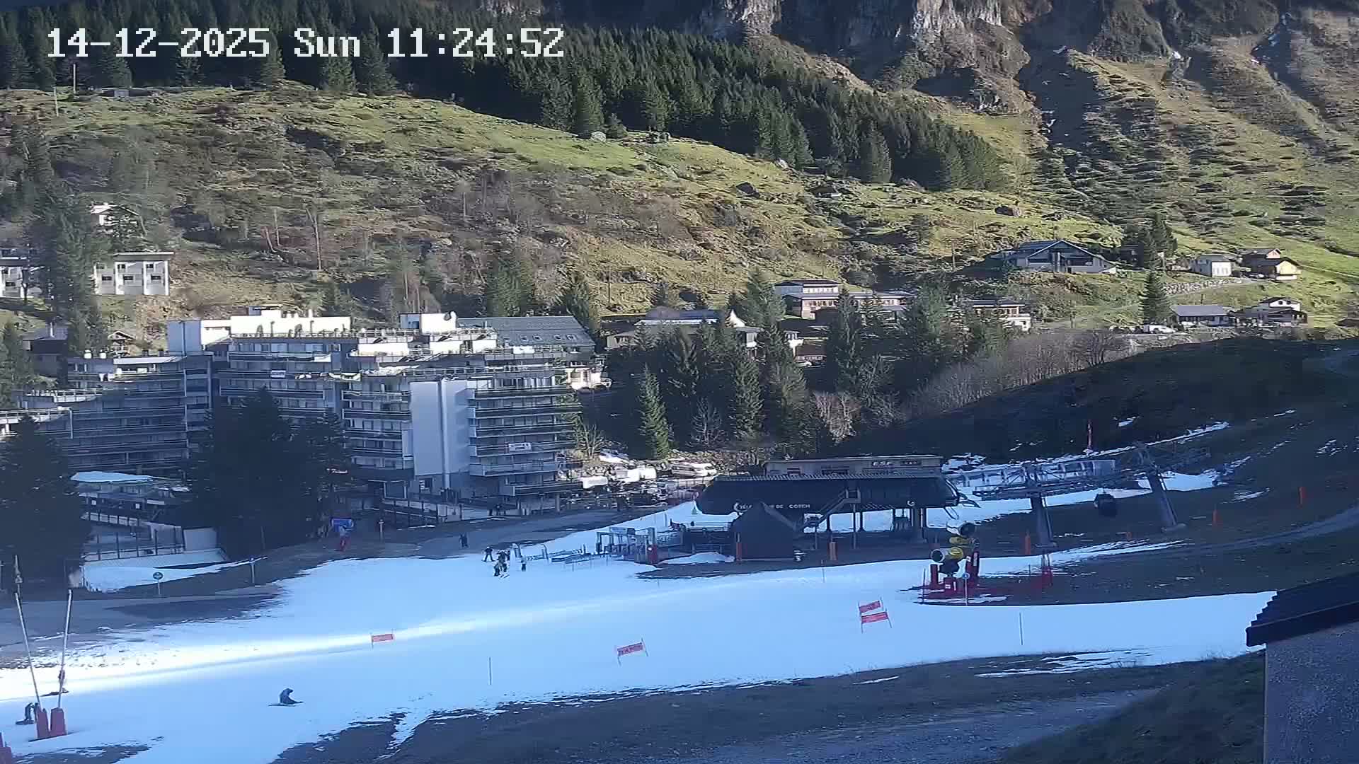 A wintry mountain landscape features a snow cannon actively making snow next to a ski lift station on a partially snow-covered slope, with multiple buildings and frosty, sparsely vegetated hillsides in the background under cold, overcast conditions.