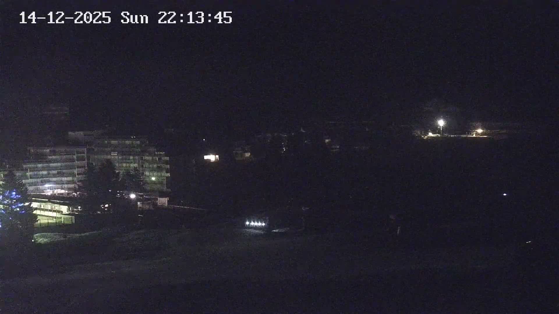 A wintry mountain landscape features a snow cannon actively making snow next to a ski lift station on a partially snow-covered slope, with multiple buildings and frosty, sparsely vegetated hillsides in the background under cold, overcast conditions.