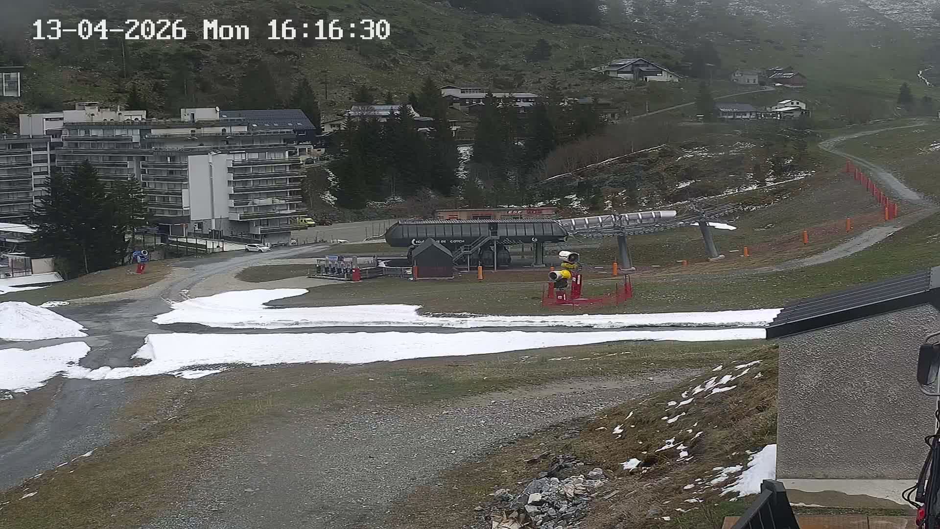 A wintry mountain landscape features a snow cannon actively making snow next to a ski lift station on a partially snow-covered slope, with multiple buildings and frosty, sparsely vegetated hillsides in the background under cold, overcast conditions.