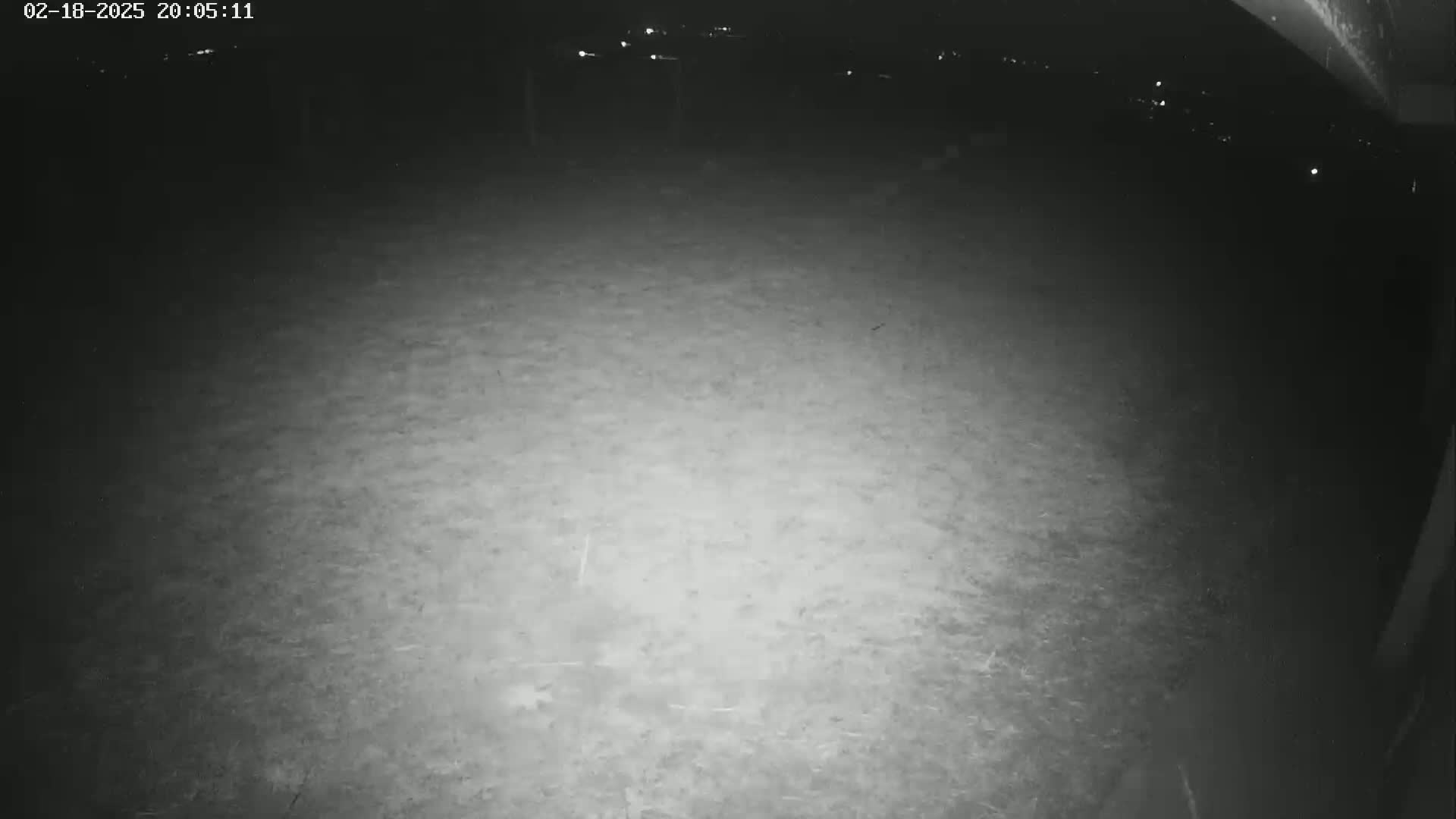 A nighttime, low-light view of a mostly empty, flat, dirt or gravel area, partially illuminated by a light source.