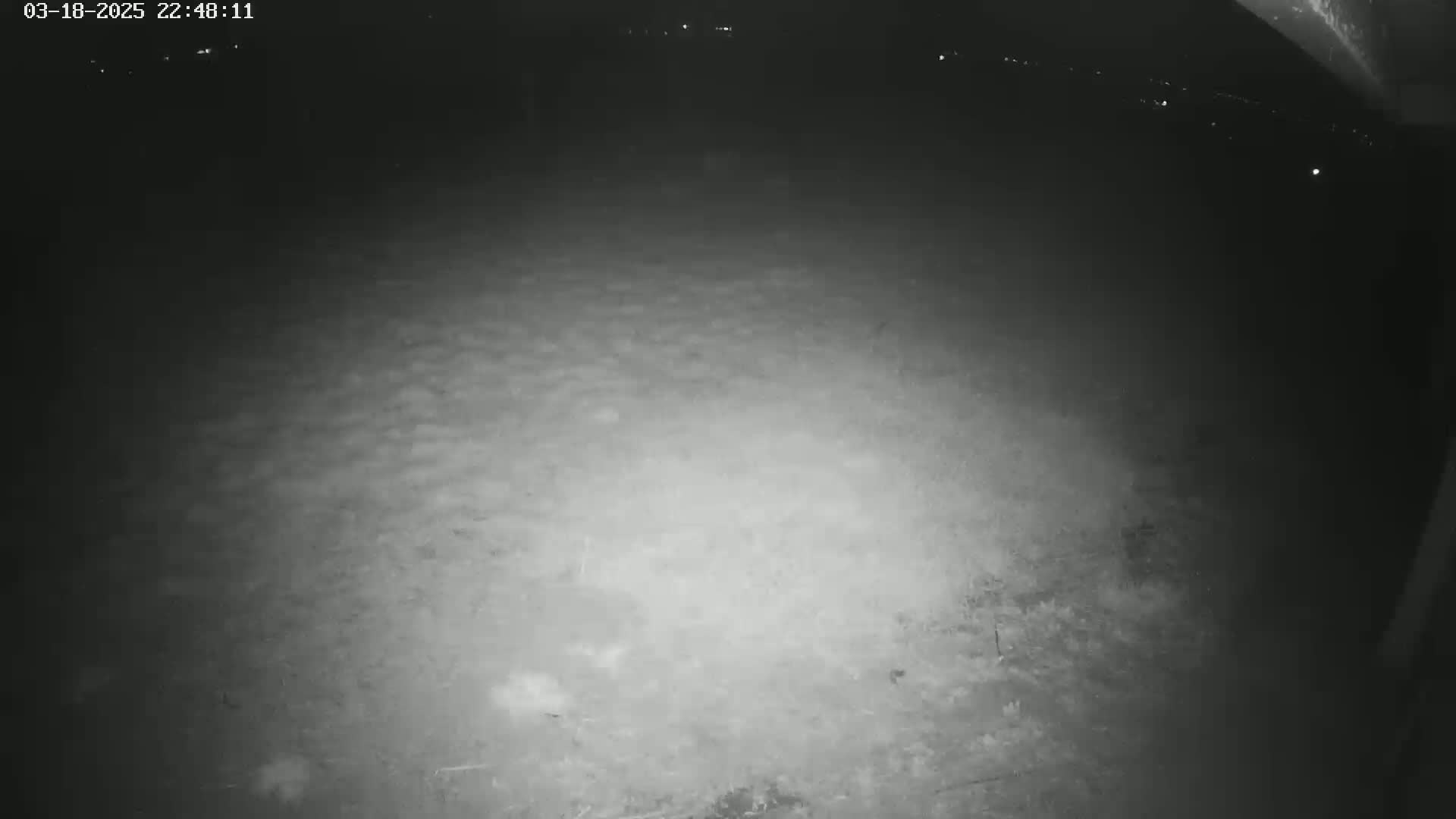 A nighttime, low-light image shows a dark, ground-level view of an uneven dirt or gravel surface, possibly taken under a covered area.