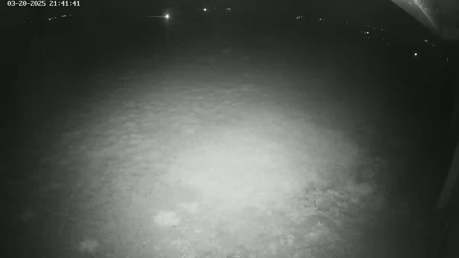 A nighttime, low-light view of a mostly empty, flat, ground-level area with distant lights visible in the background.