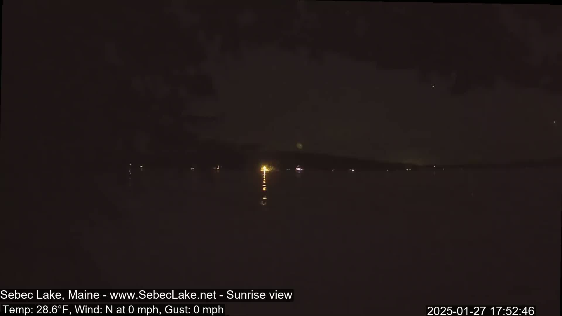 A nighttime view of a calm lake with sparse lights on the far shore under a dark sky.