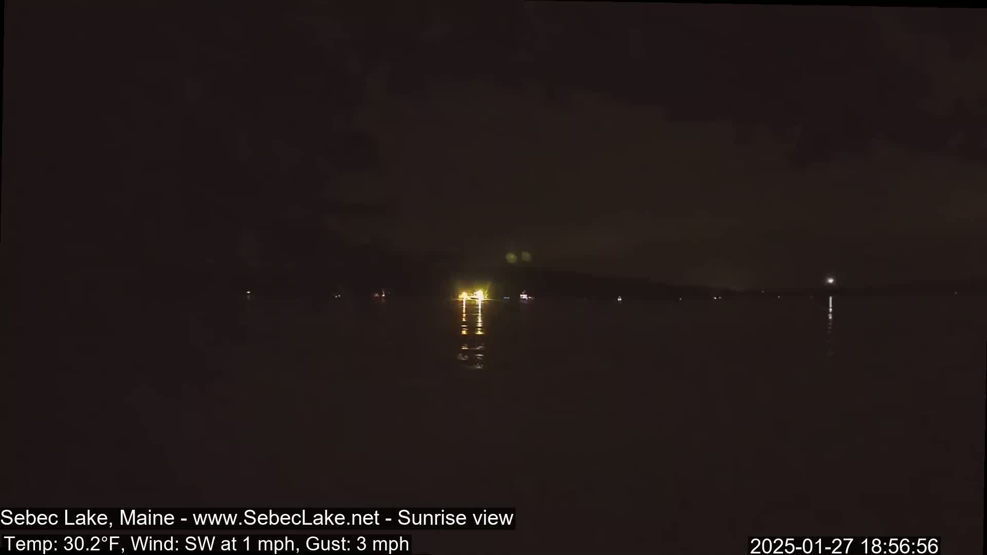 A dark nighttime view of a lake with faint lights reflecting on the water, under a mostly clear sky with a southwest wind.