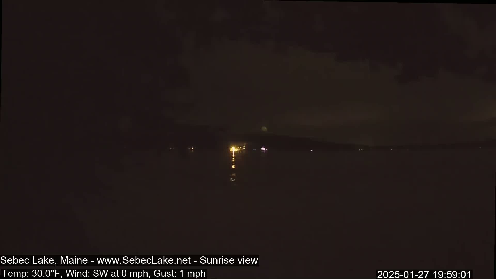A nighttime view of a dark lake with a few distant lights on the far shore under a mostly cloudy sky.