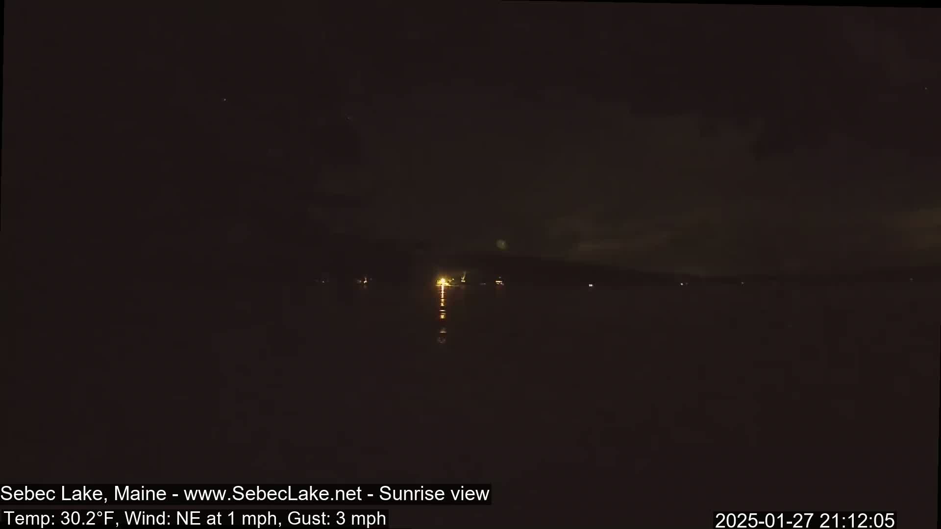 A dark, nighttime scene shows a body of water reflecting a few distant lights under a mostly clear sky with a slight northeast wind.