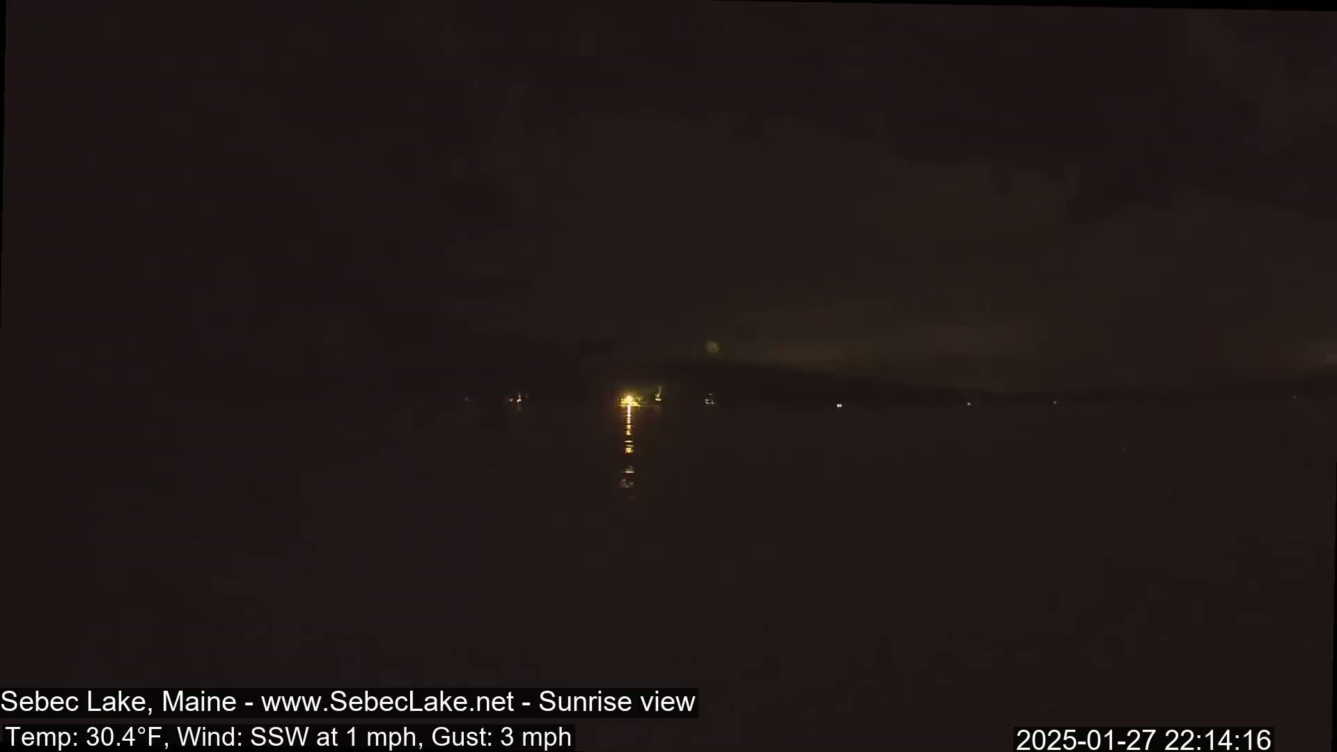 A dark nighttime view of a lake with faint lights on the far shore and a slight reflection on the water under a mostly clear sky, with a 1 mph wind.