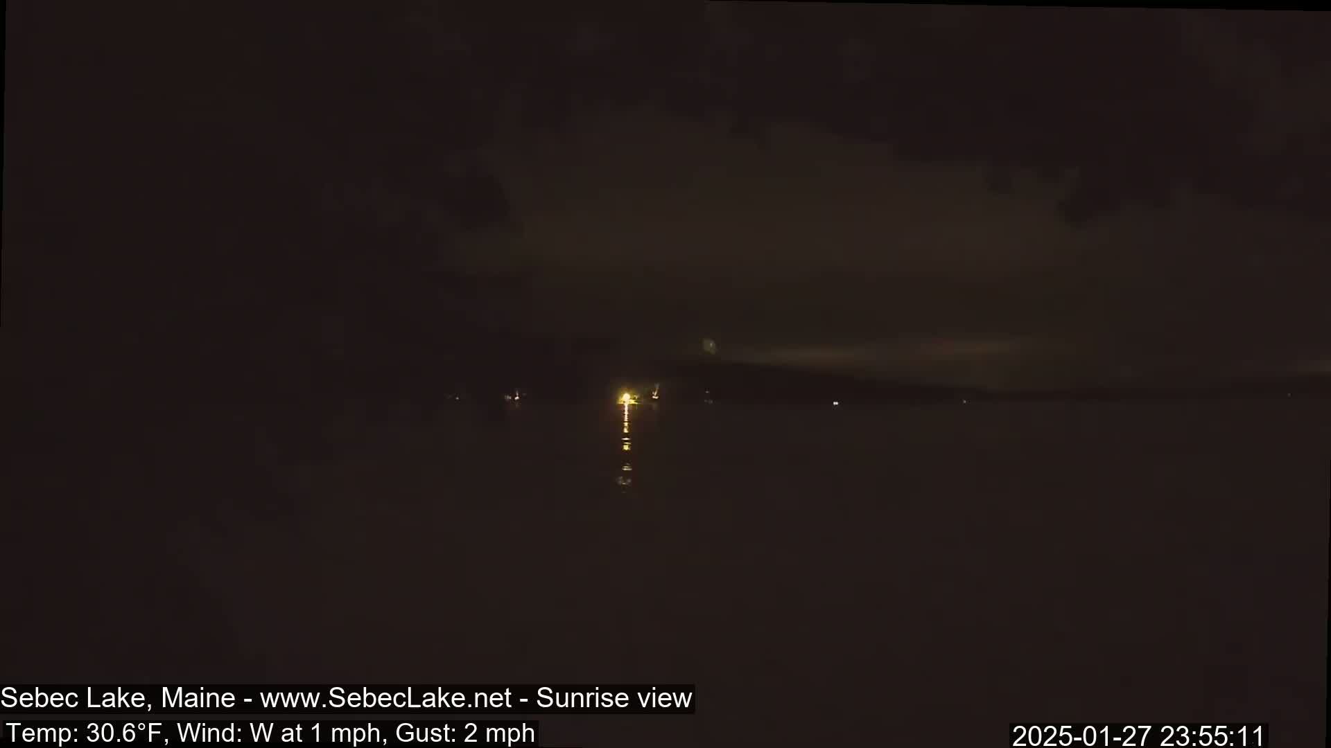 A dark, nighttime view of a lake with faint lights on the far shore and a calm, westerly wind.