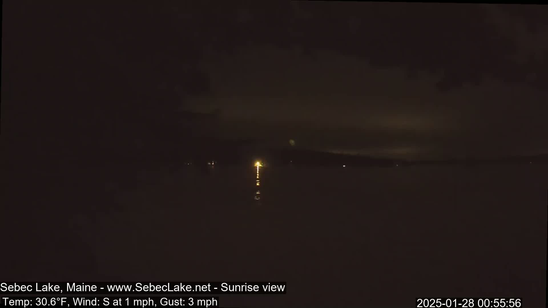 A dark, nighttime view of a calm lake with a single, distant light source reflecting on the water under a mostly cloudy sky.