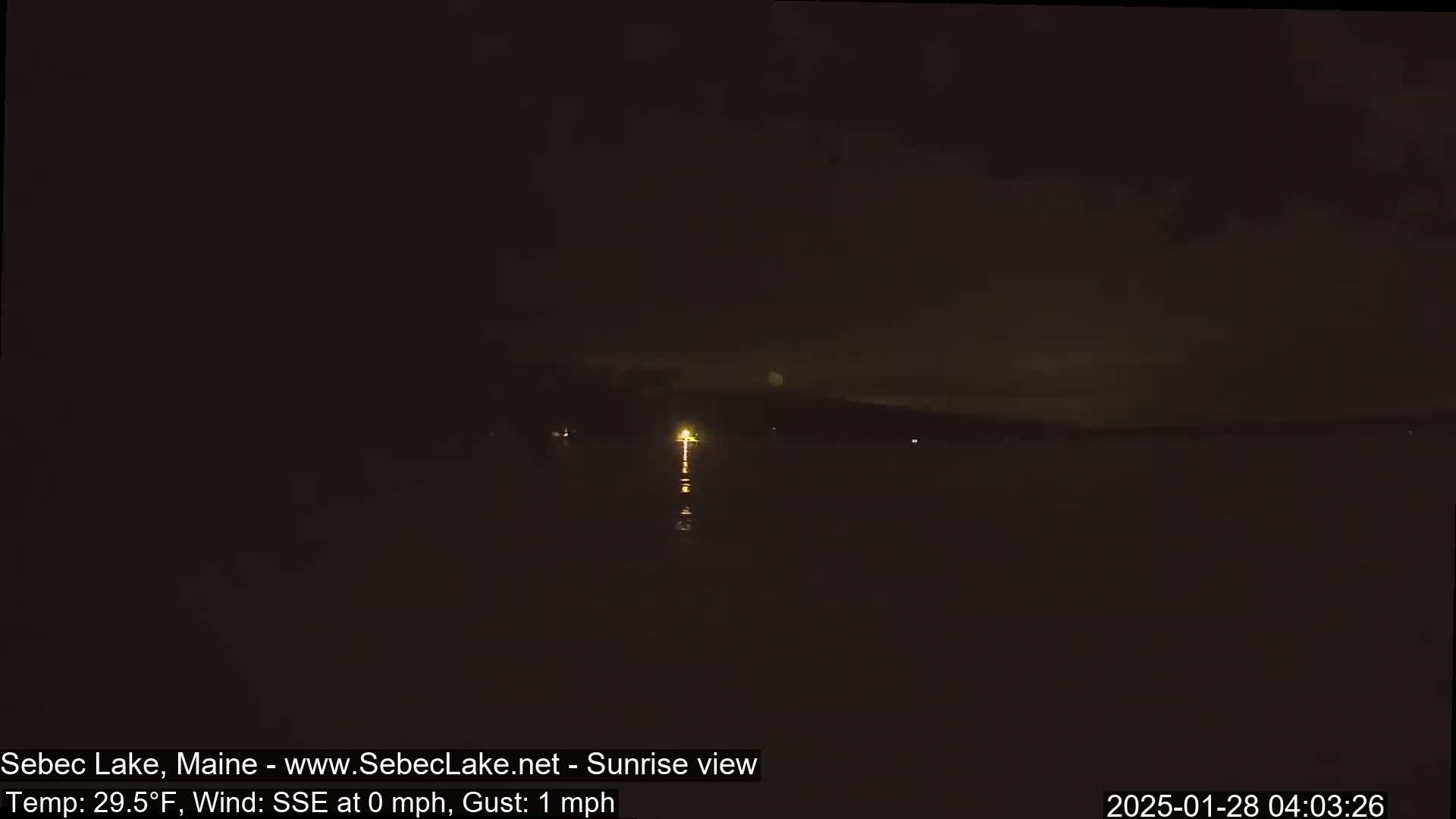 A dark, nighttime view of a lake shows a faint light on the far shore, with calm water reflecting the light under a mostly clear, dark sky.
