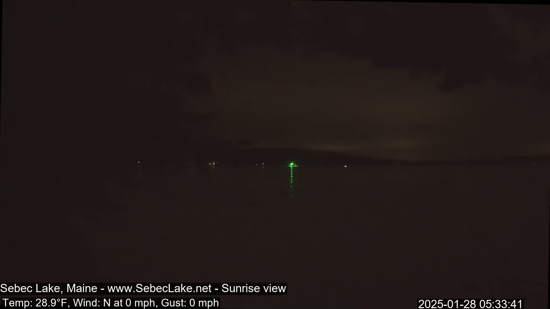 A dark nighttime view of a lake with a single green light visible on the far shore under a mostly clear sky.