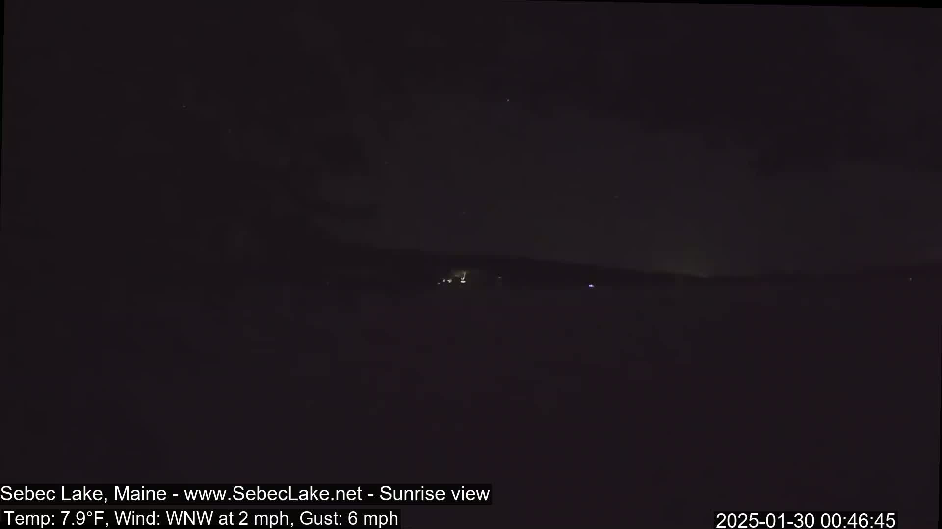 A dark, nighttime landscape shows a distant, dimly lit shoreline under a mostly clear sky with a slight wind.
