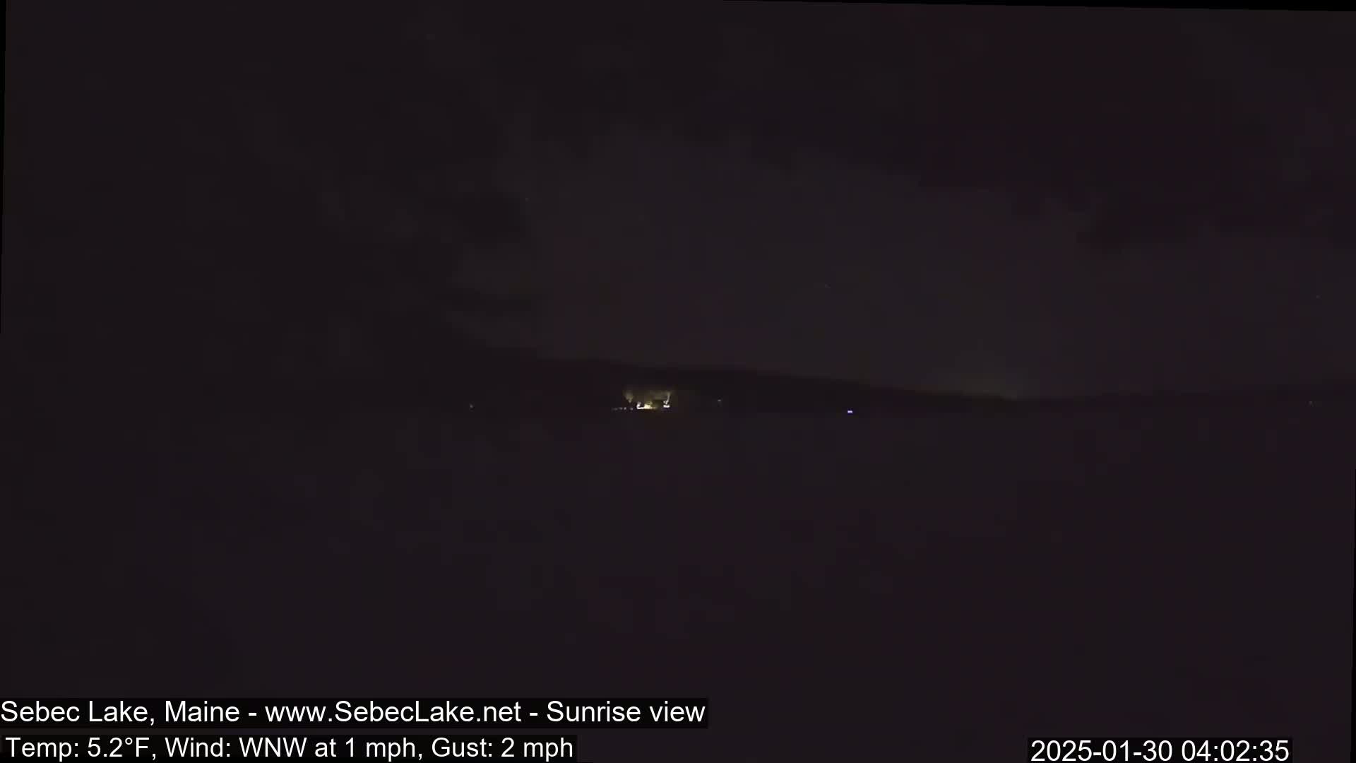 A dark, nighttime view of a lake with a faintly illuminated shoreline in the distance, under a mostly clear sky.