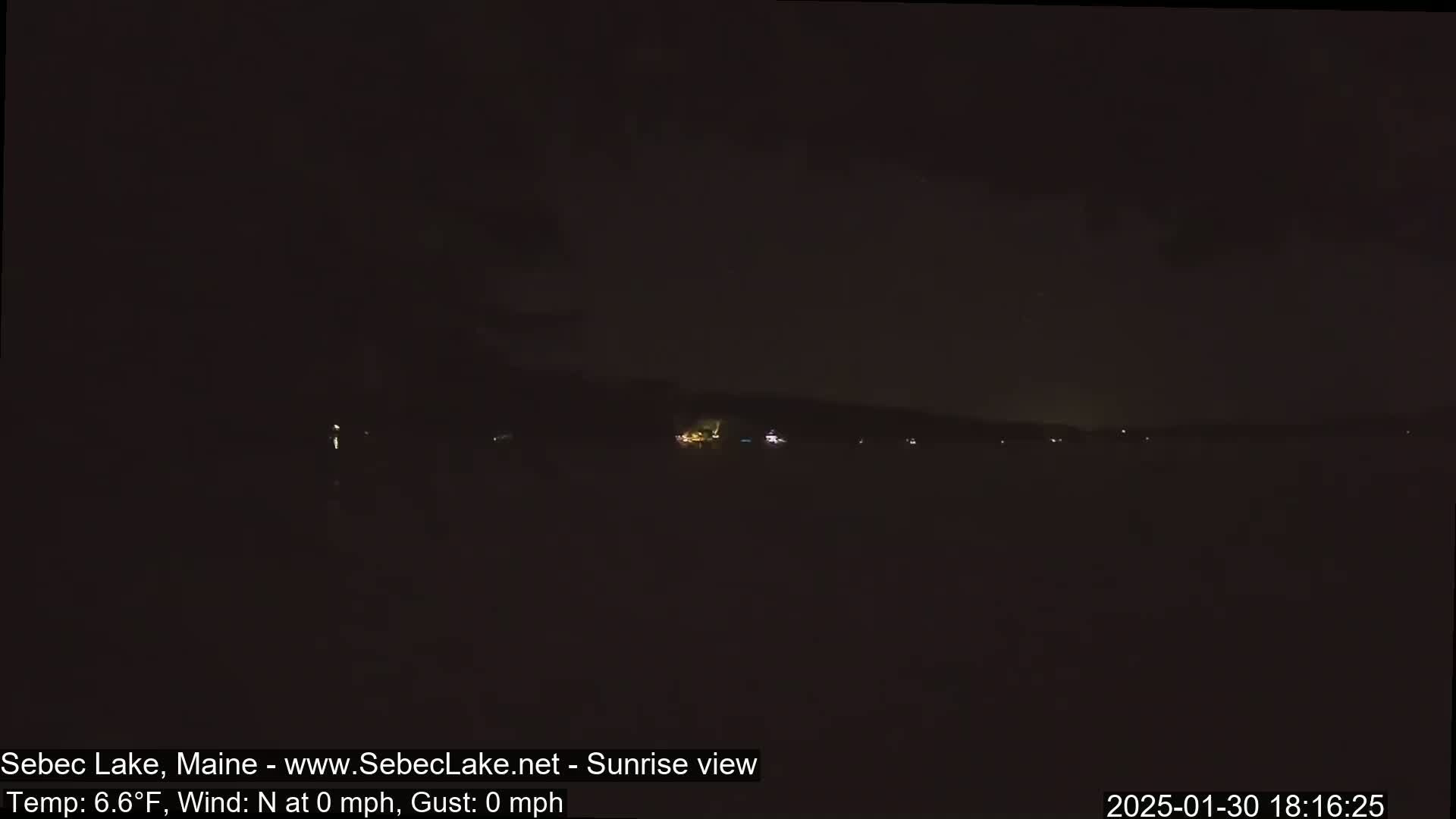 A dark, nighttime view of a calm lake with a few distant lights visible on the far shore under a mostly clear sky.