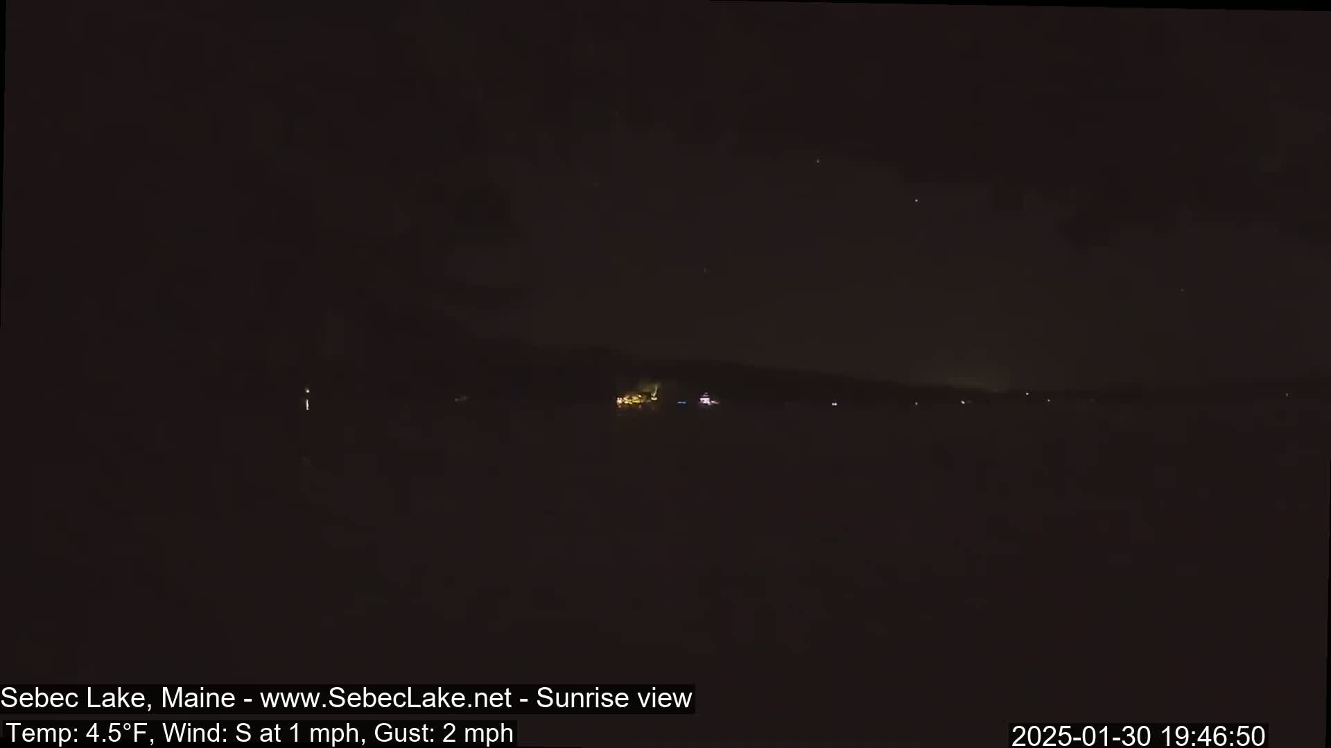 A dark, nighttime view of a lake with a distant shoreline showing a few faint lights, under a clear sky with a southerly wind.