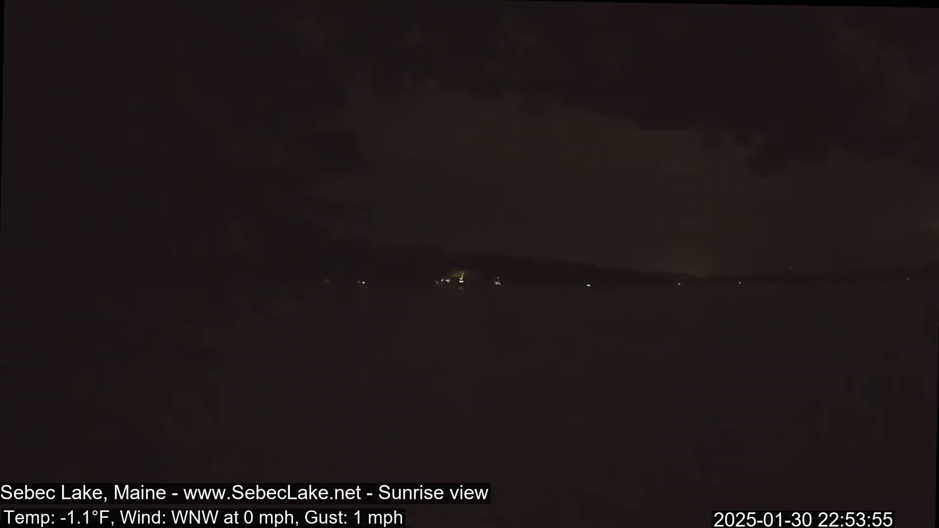A dark, nighttime view of a lake with a distant, faintly lit shoreline under a cloudy sky.