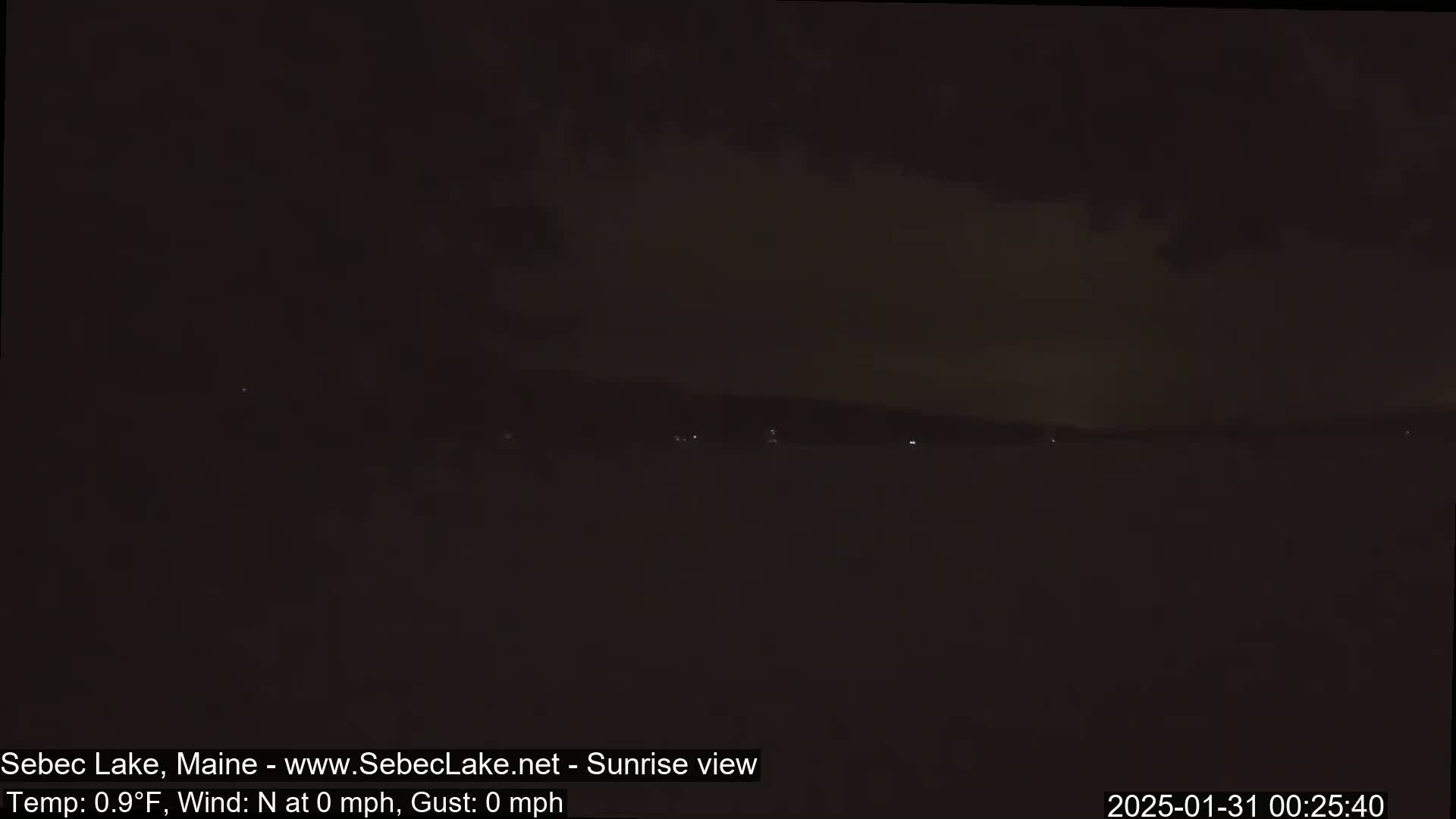 A dark, nighttime view of a calm lake with a distant, faintly lit shoreline under a mostly cloudy sky.