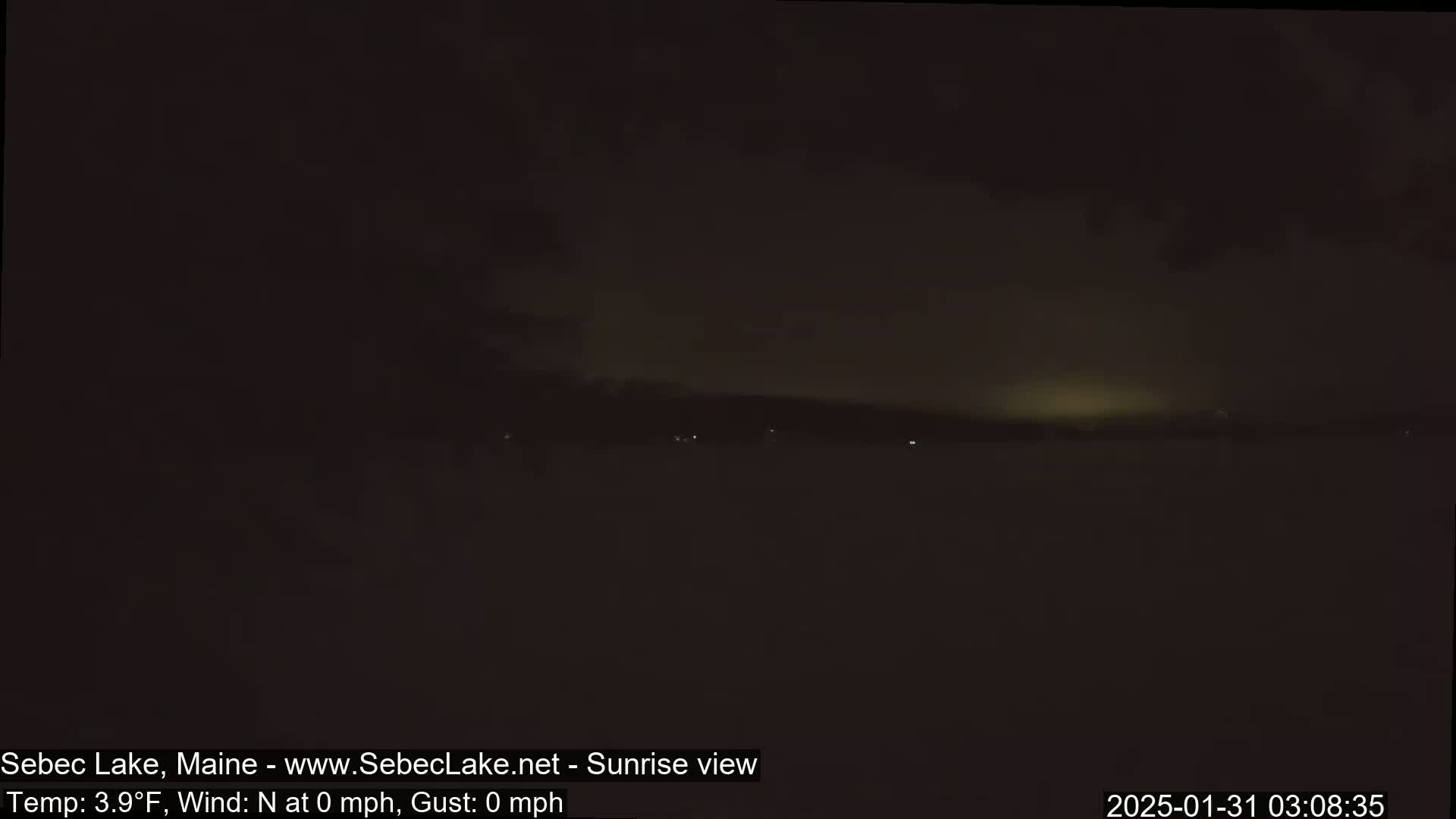 A dark, pre-dawn view of a calm lake under a mostly cloudy sky.